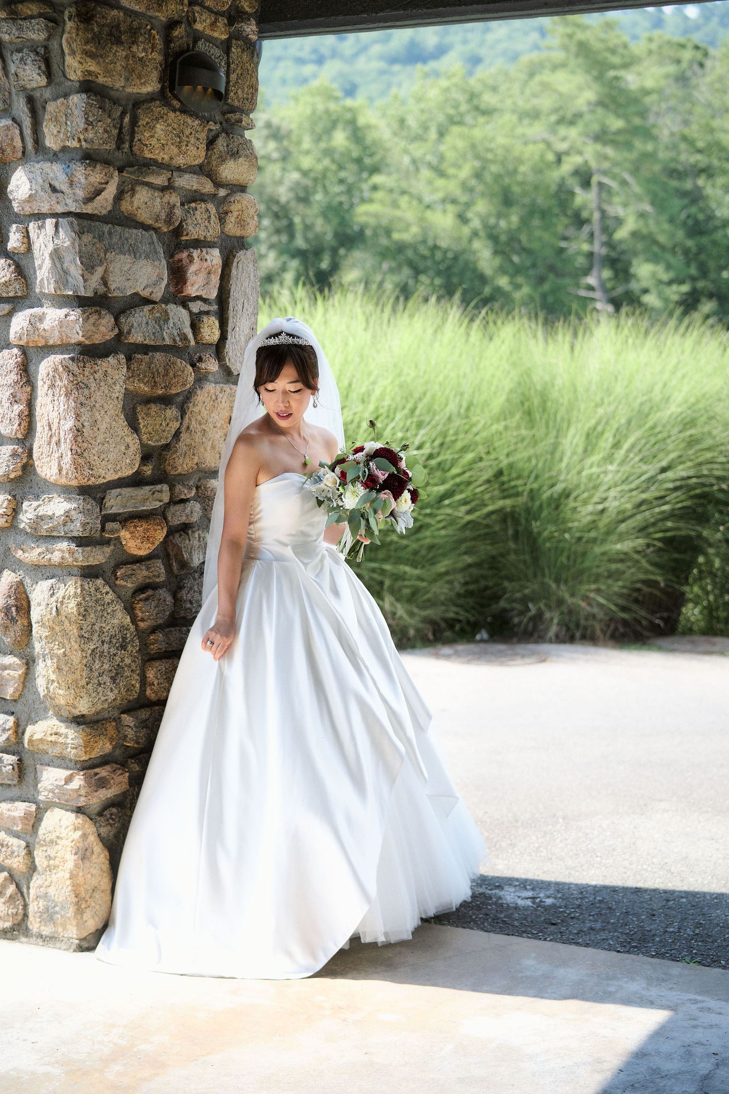 A bride in a classic white ballgown holding a bouquet poses by a rustic stone pillar.