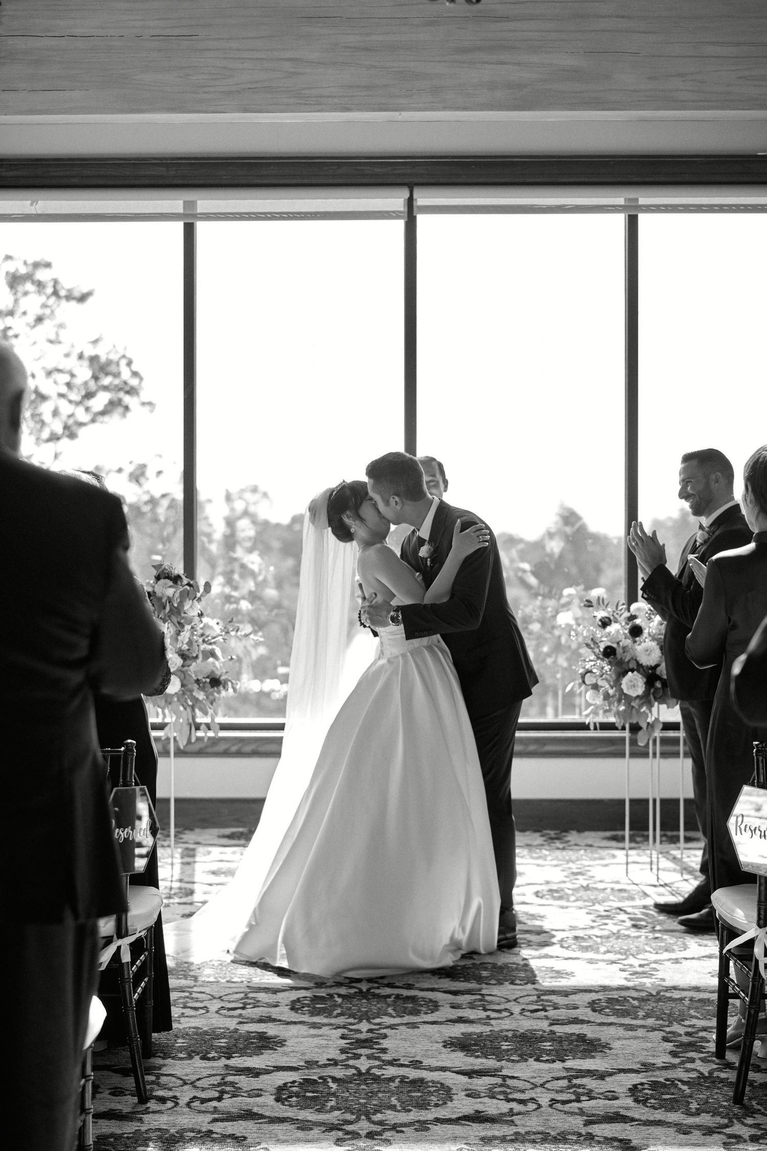 Black and white wedding ceremony photo with dramatic window lighting and floral decorations.