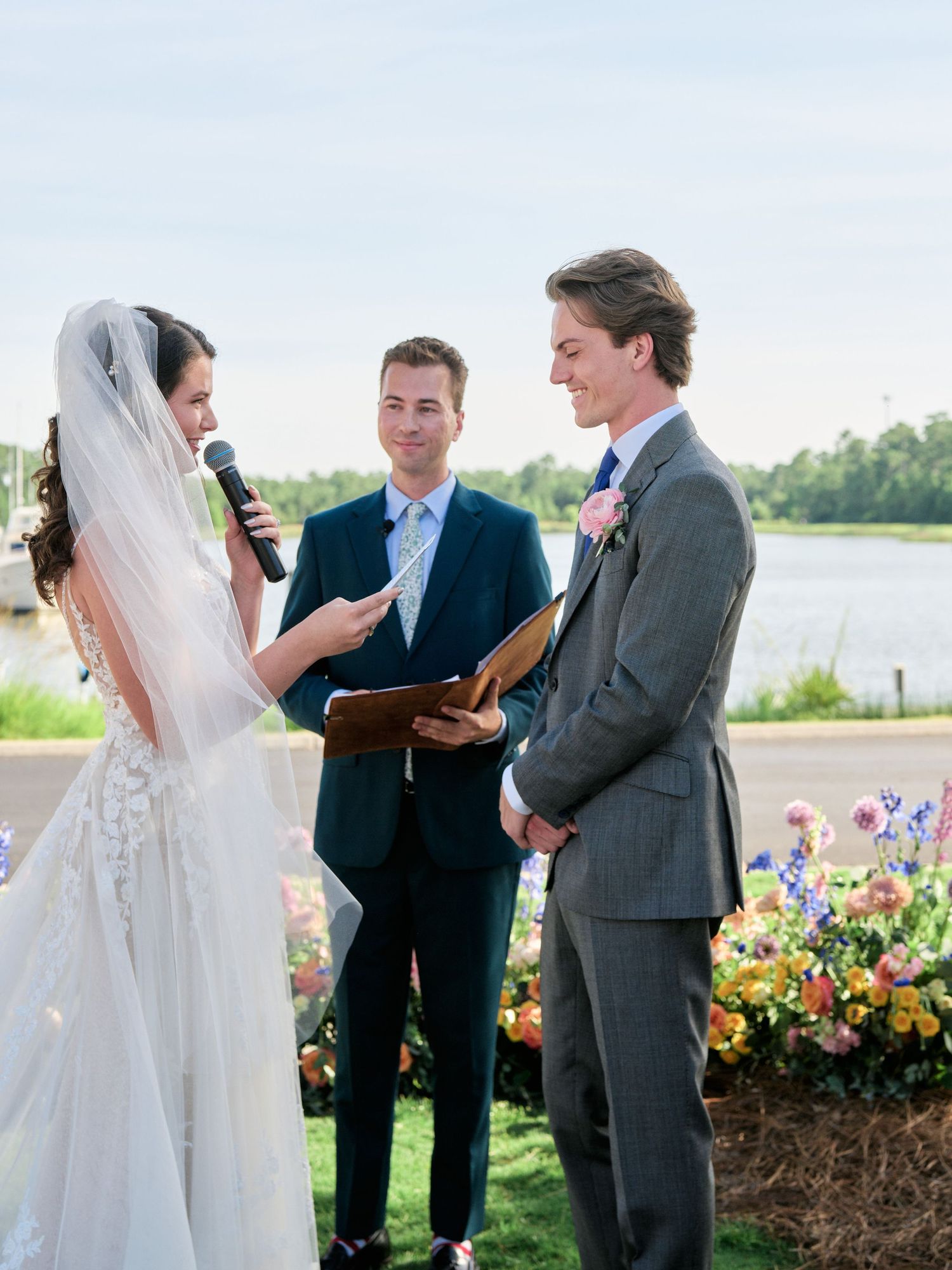 An outdoor wedding ceremony takes place by a waterfront with colorful flower arrangements adorning the venue.