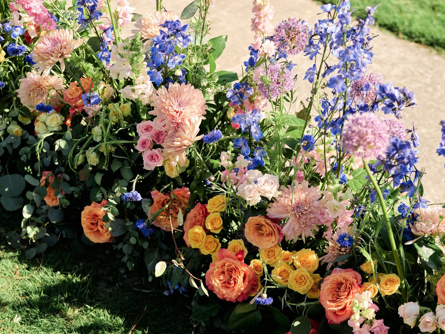 A colorful garden border with pink carnations, peach roses, and blue delphinium flowers blooming in summer sunshine.