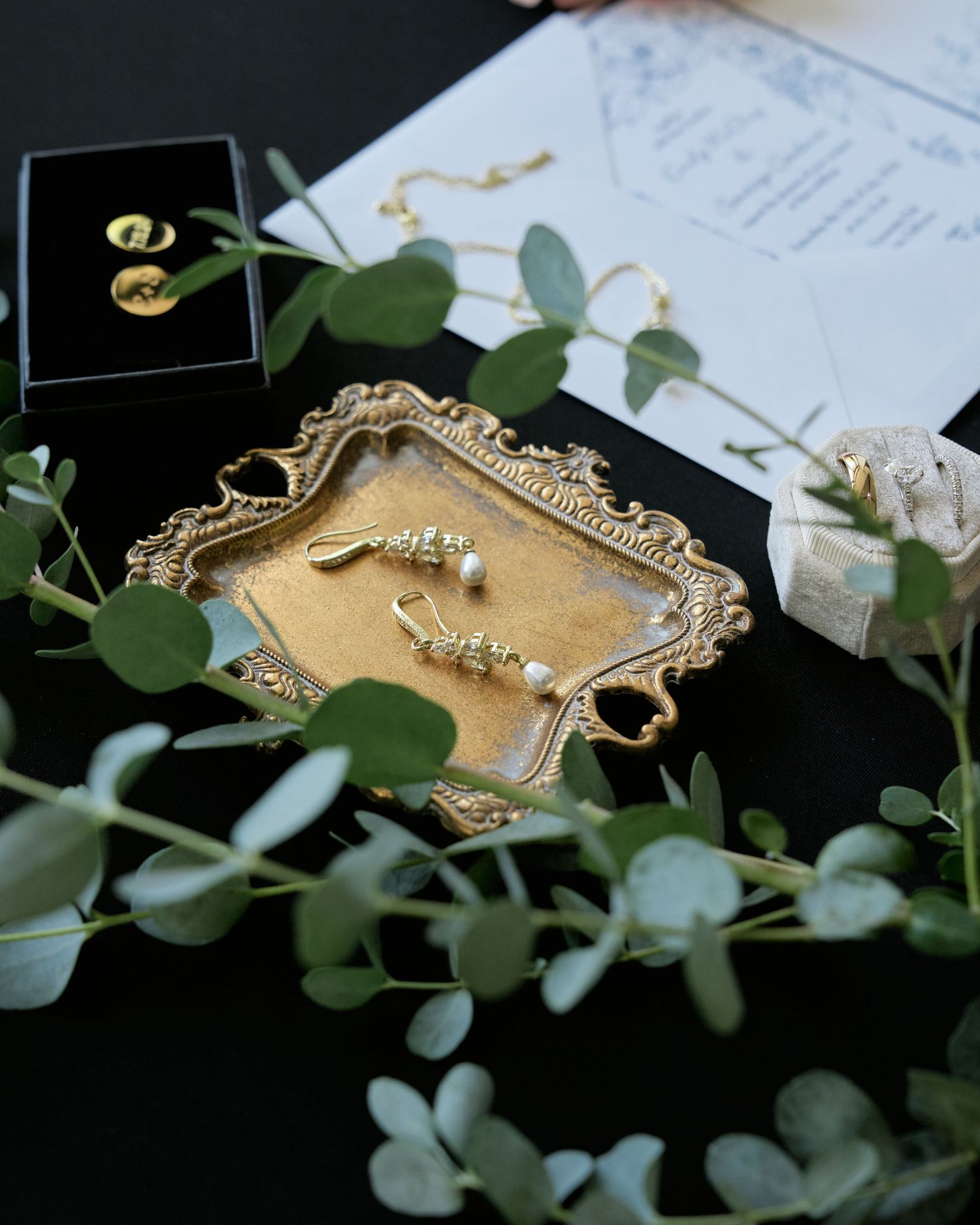 Wedding jewelry displayed on ornate gold tray with eucalyptus greenery and invitation details.