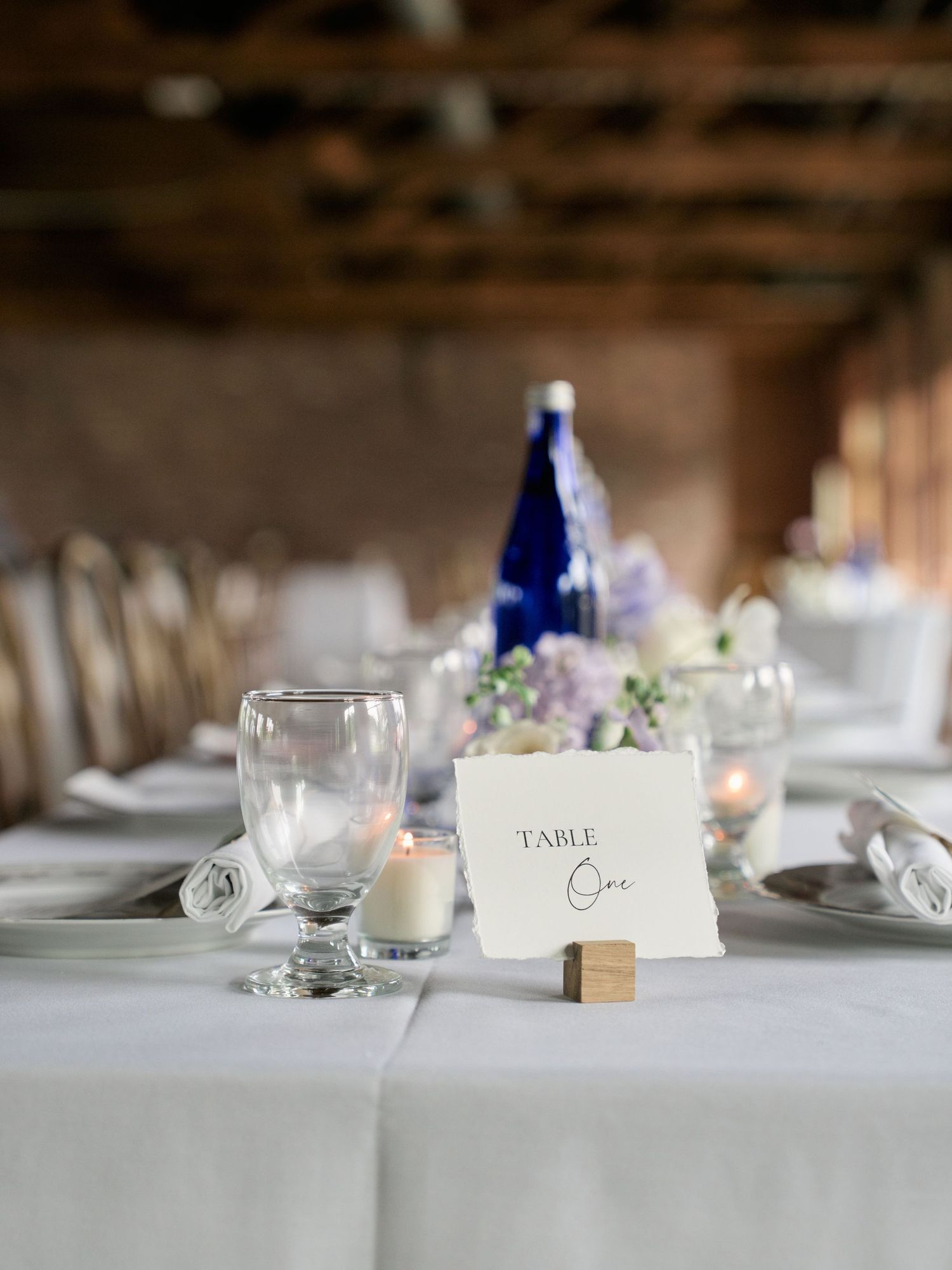 Wedding reception table place card and elegant table setting with white linens, wine glasses, and blue bottle decor.