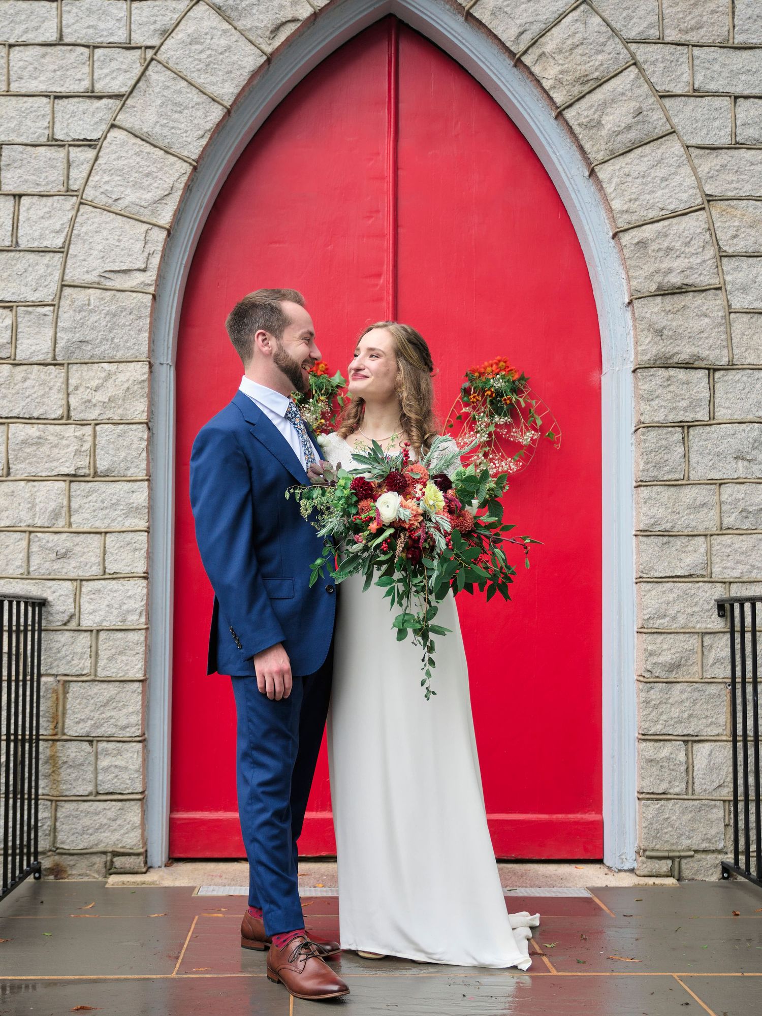 A couple shares an intimate moment in front of a vibrant red church door while holding a lush floral bouquet.