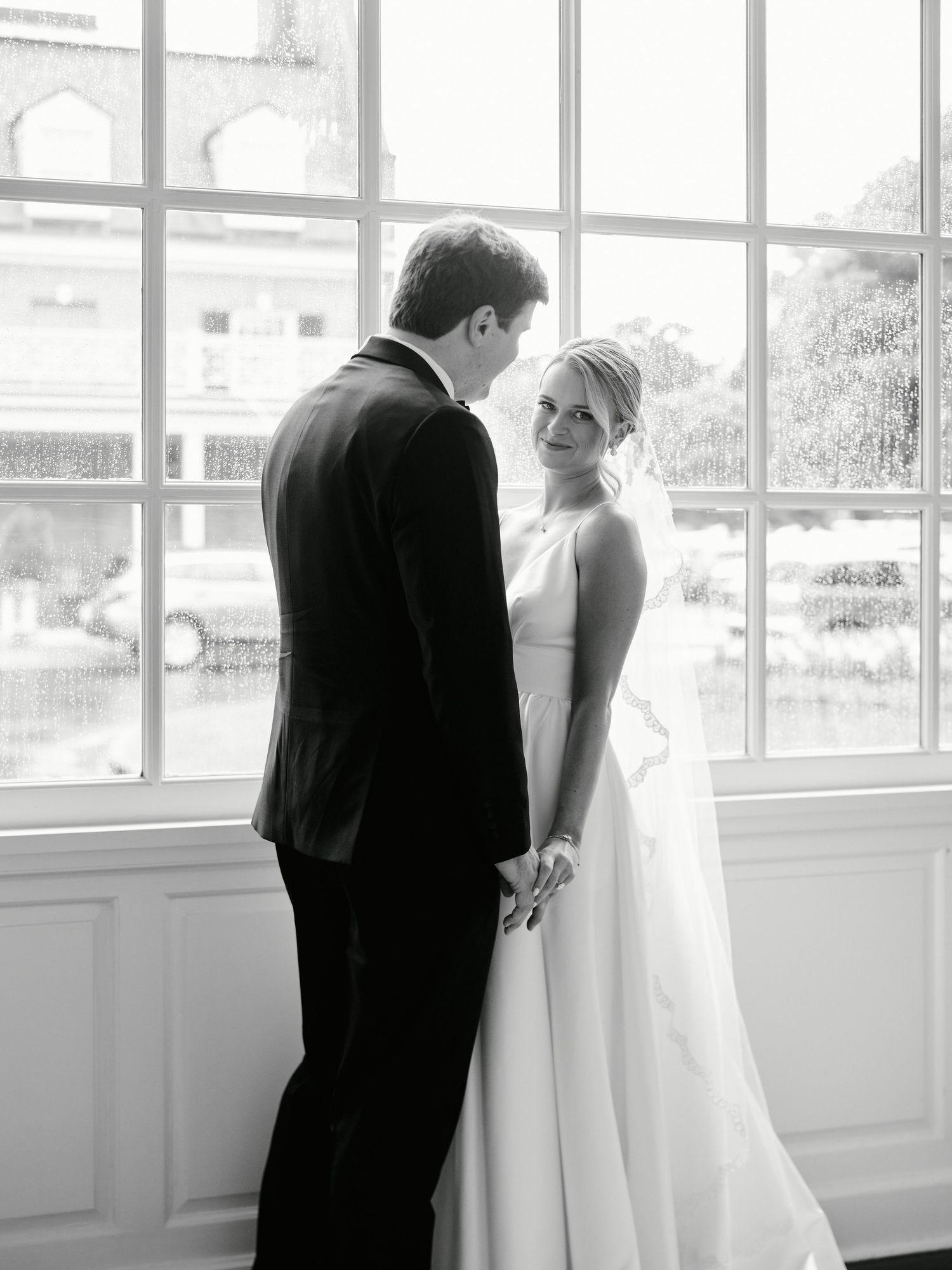 Black and white portrait of couple by large windows in classic wedding venue.