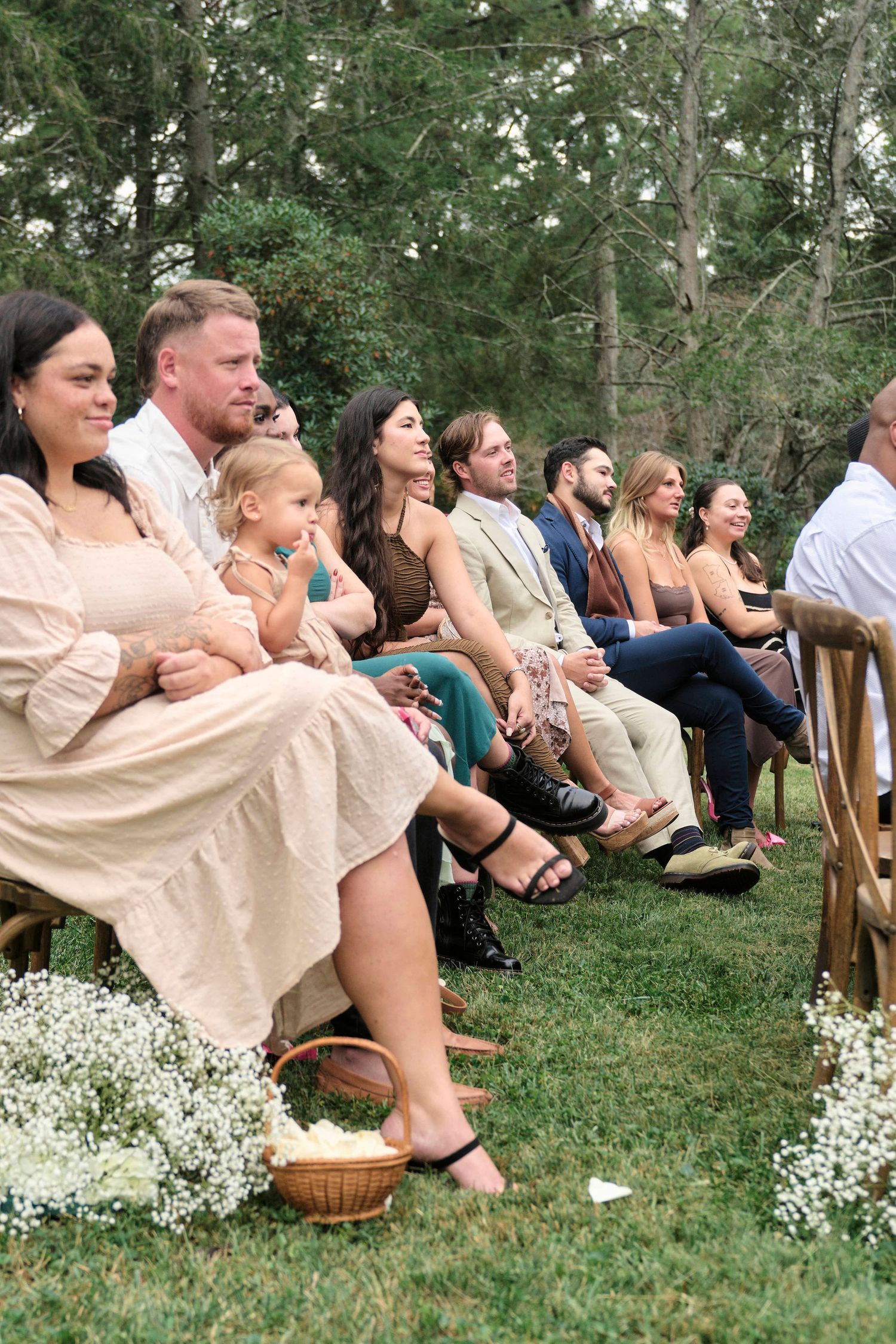 Wedding guests seated on wooden chairs at outdoor ceremony with white floral decorations.