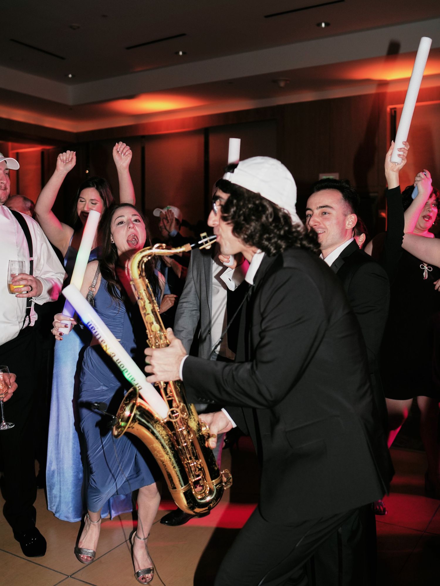 Musicians and party guests dance with light sabers and play saxophone at a festive indoor celebration with red lighting.
