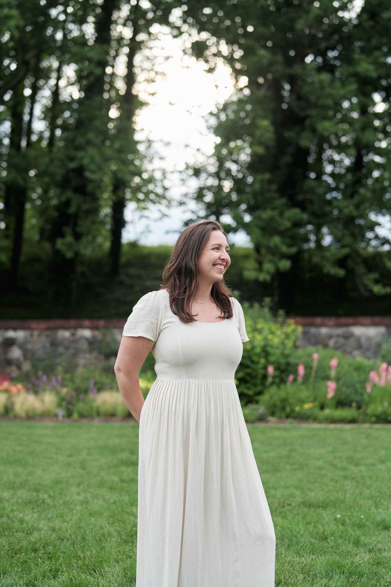 A person in a flowing white dress smiles while standing in a garden setting at dusk.