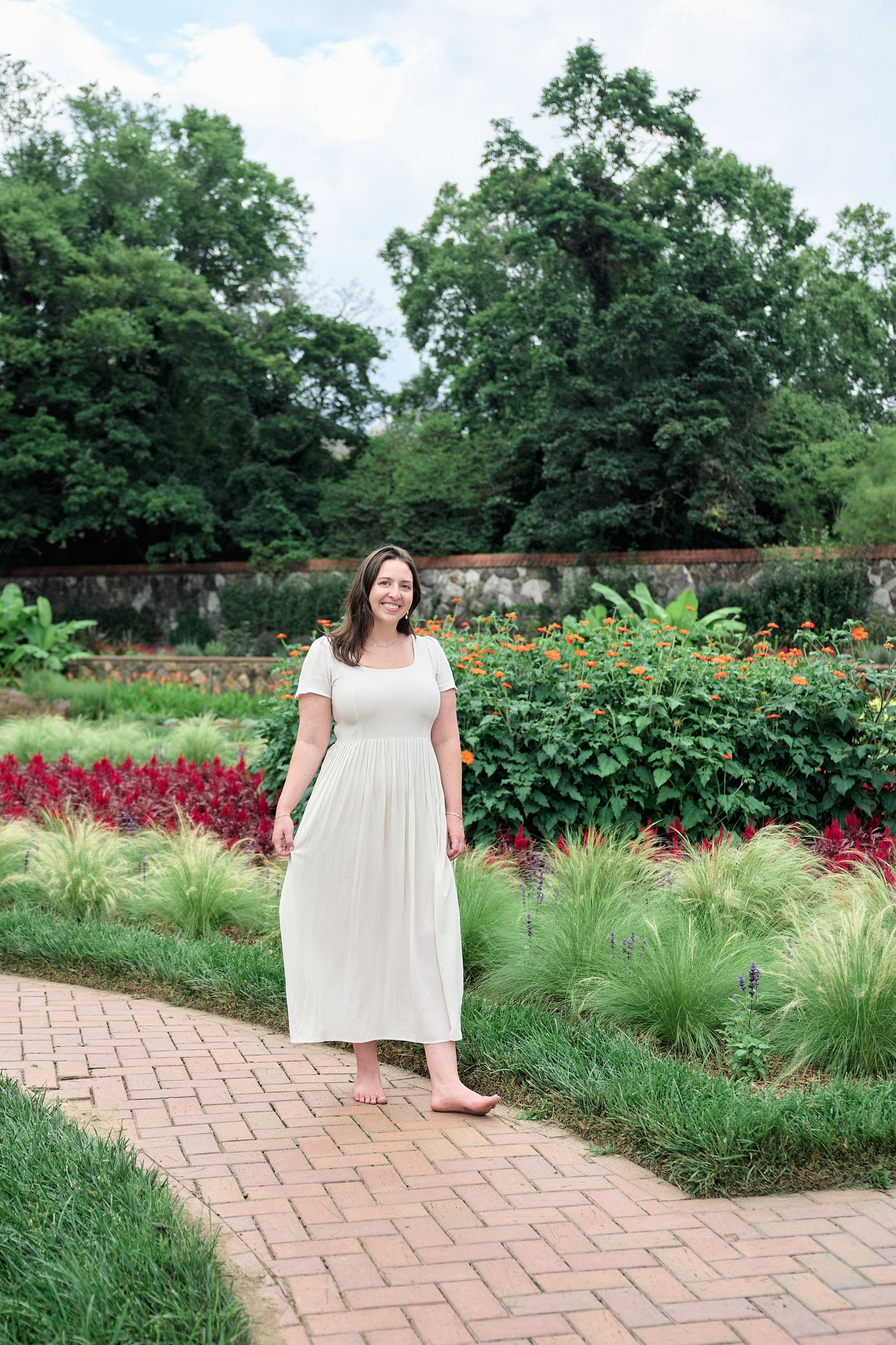 Woman in cream dress walks along brick pathway in flowering garden.