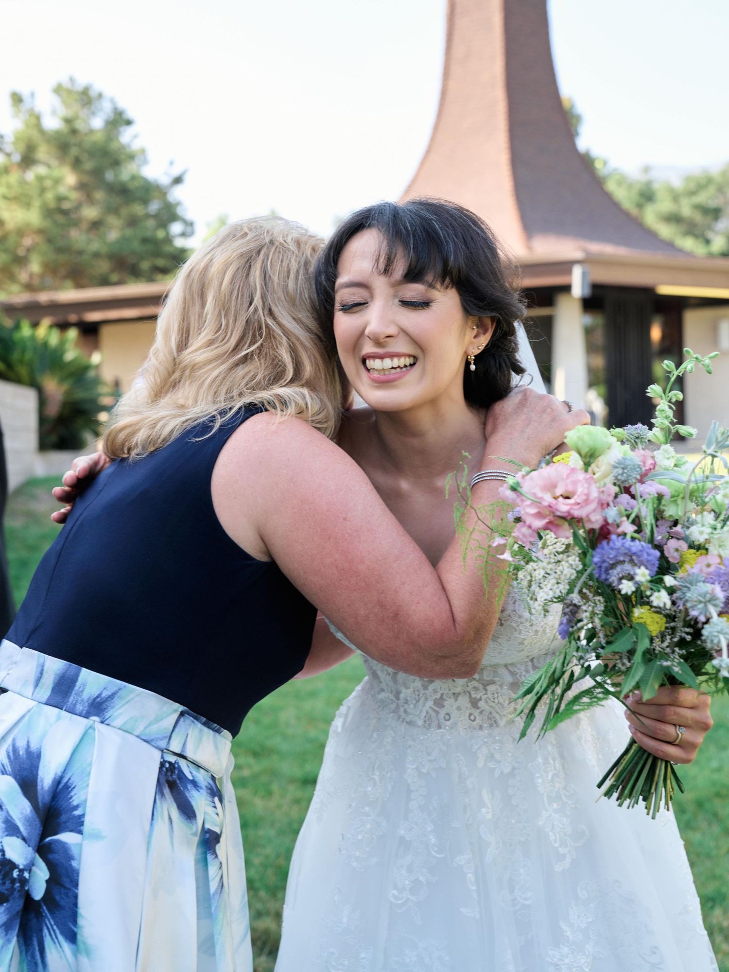 Two people share an emotional hug at an outdoor wedding while holding a colorful bouquet.