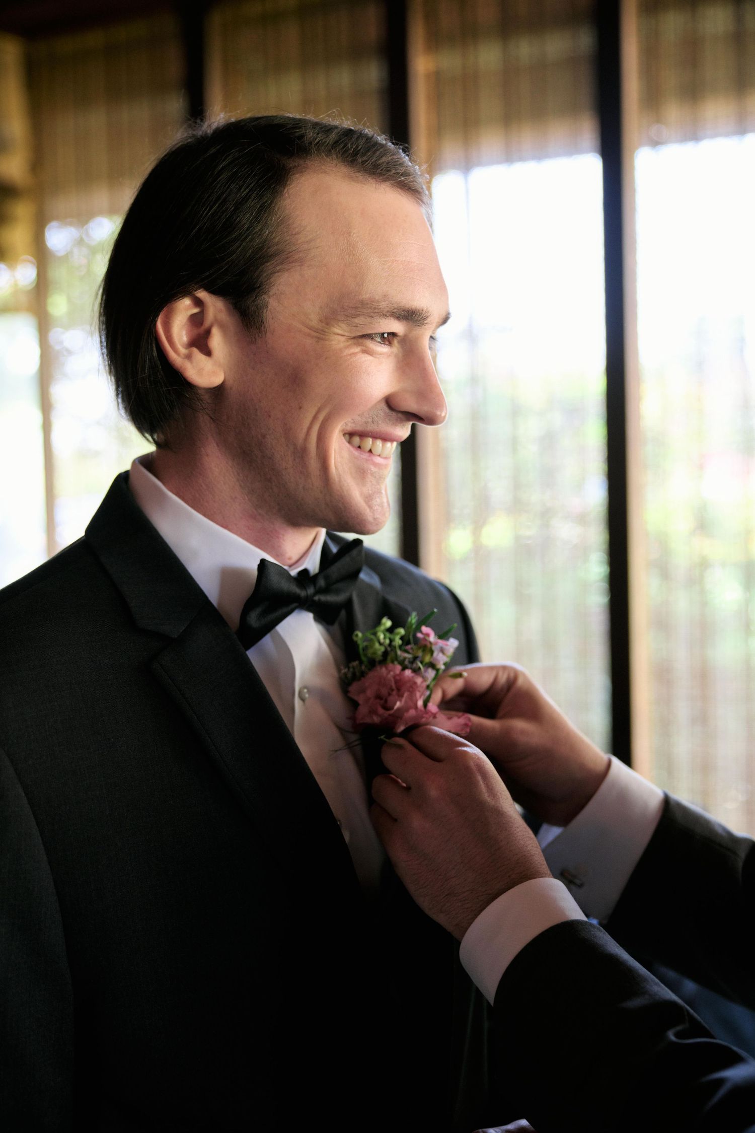 A groom in a black tuxedo smiles while having a boutonniere pinned to his lapel during wedding preparations.
