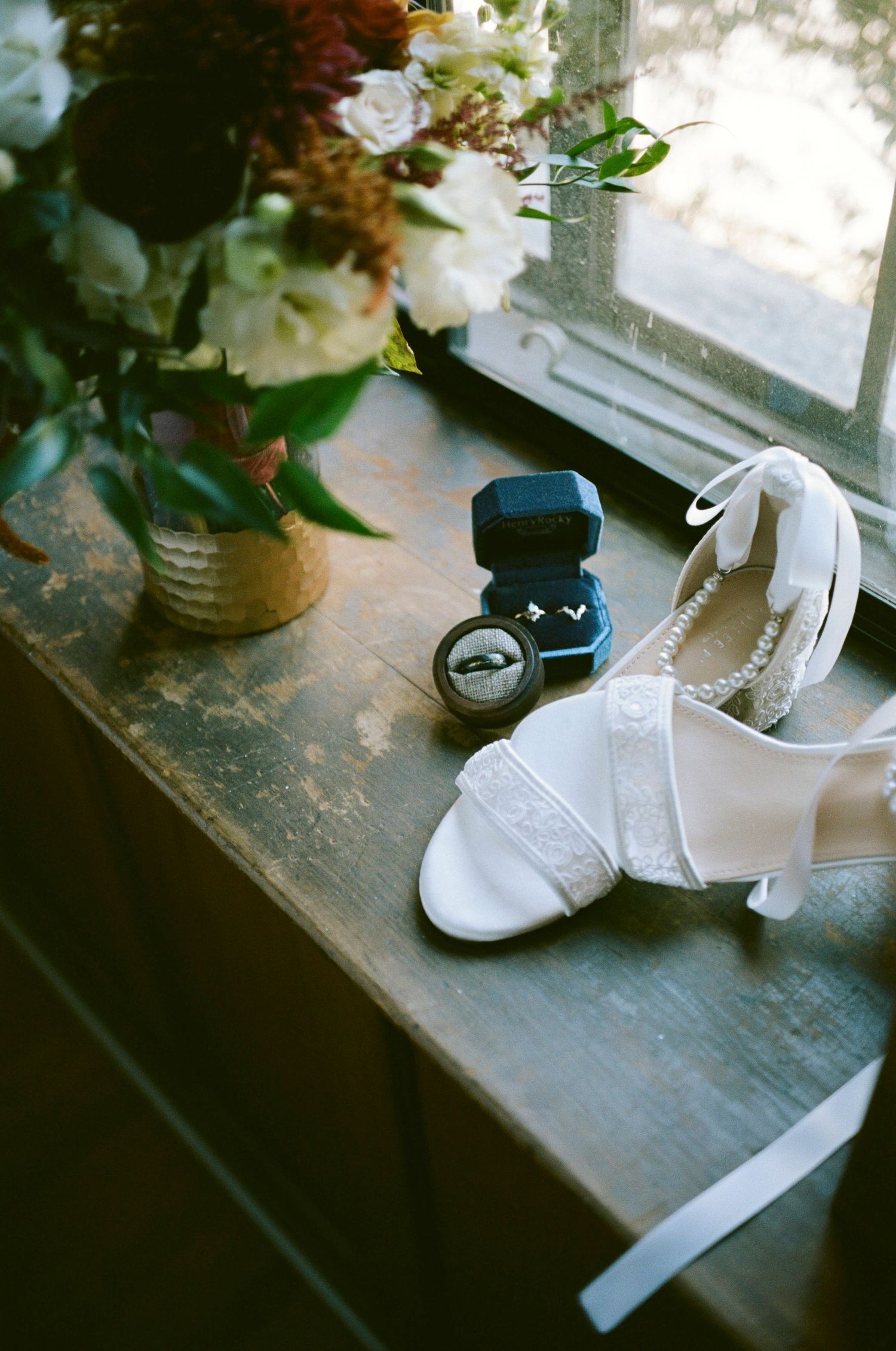 White bridal shoes and ring box displayed on rustic wooden windowsill with floral arrangement.