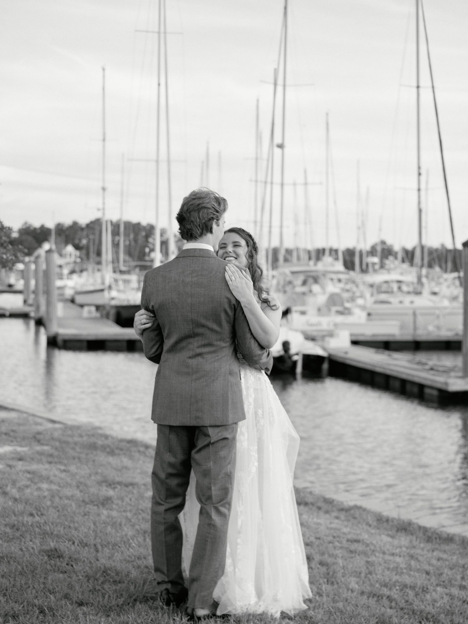A romantic black and white wedding moment captured at a marina with sailboats in the background.