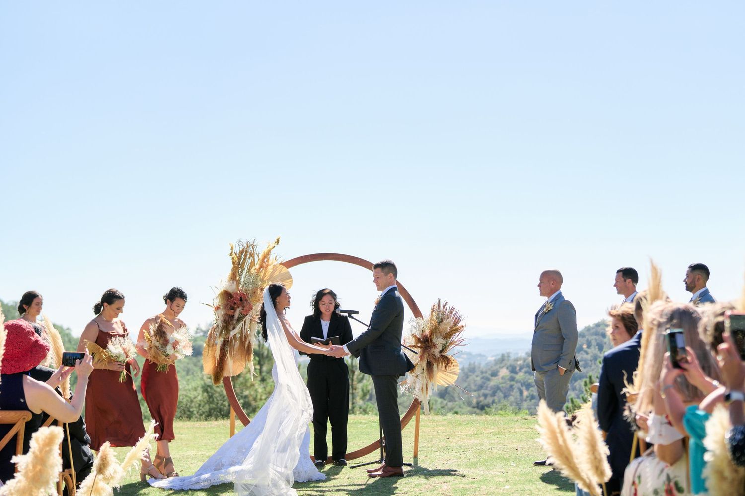 Wide view of an outdoor wedding ceremony with guests seated on wooden chairs beneath sunny skies.