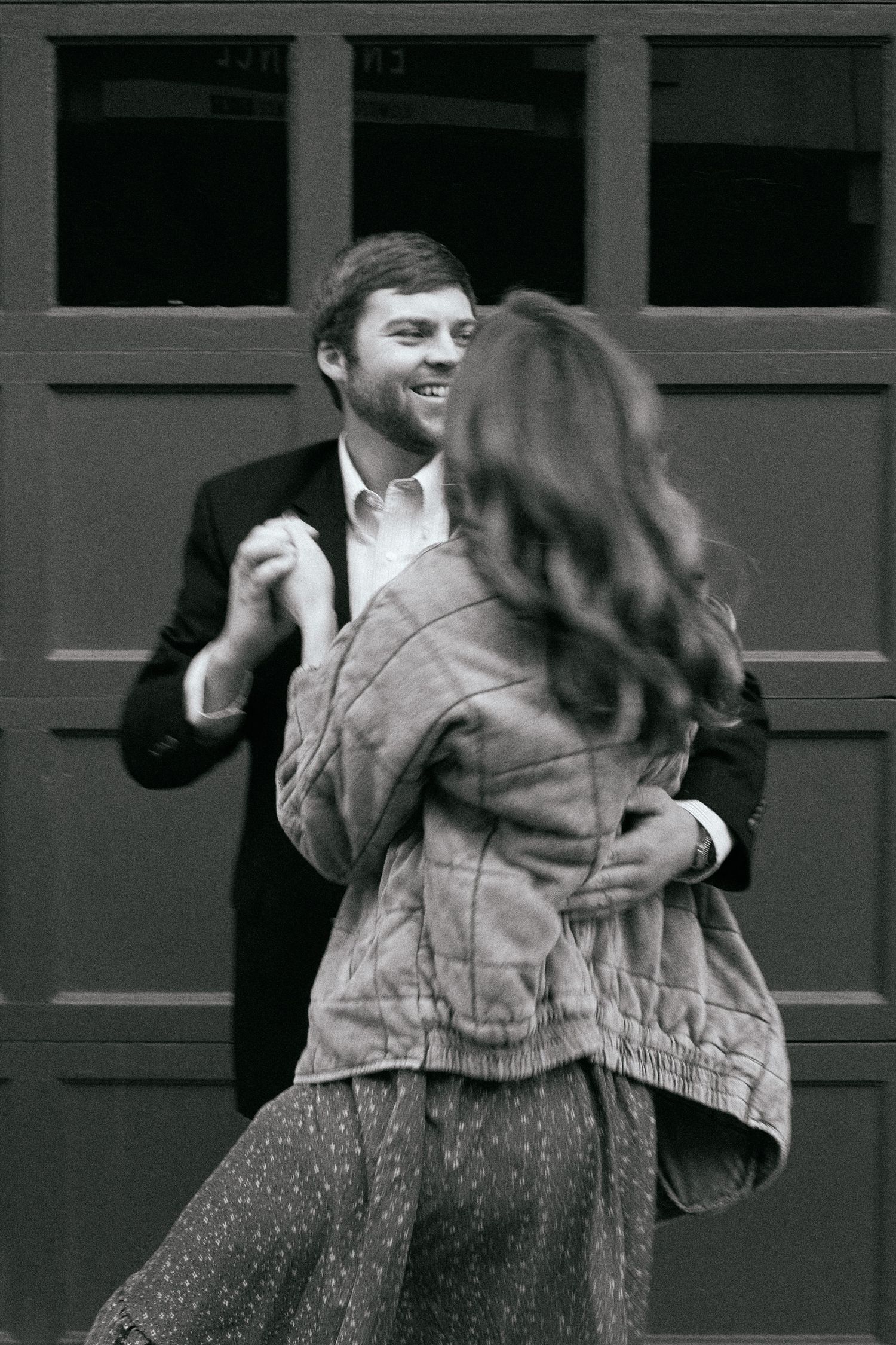 A black and white photo capturing a couple dancing and laughing together against a dark wall.