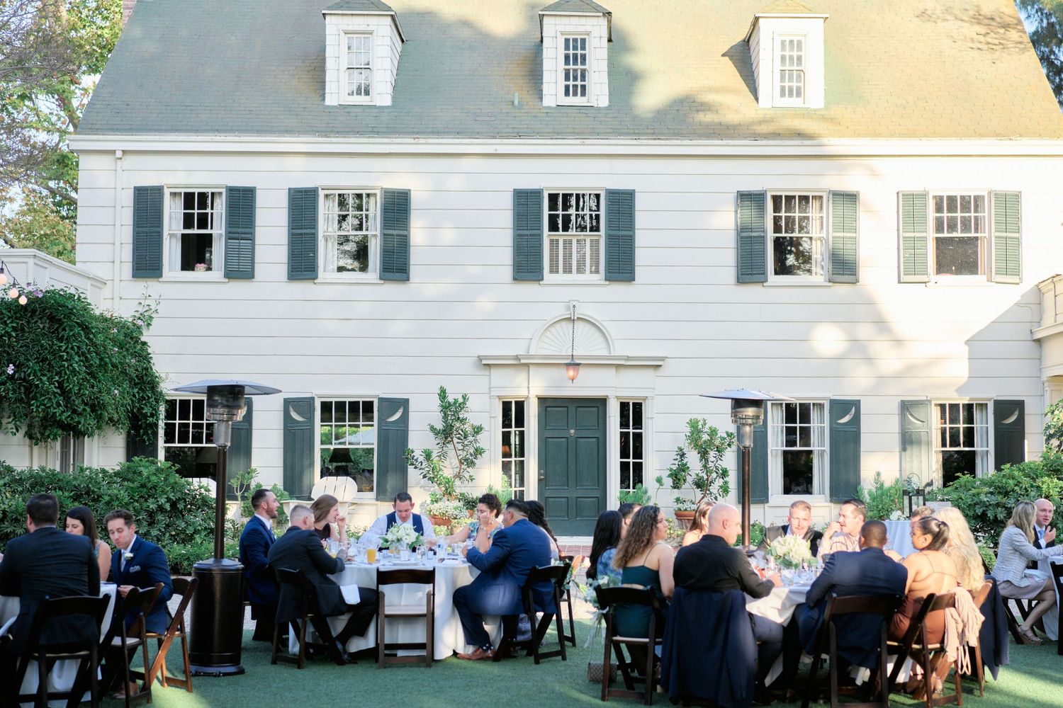 Elegant outdoor wedding reception at a classic white colonial-style venue with guests dining on the lawn.