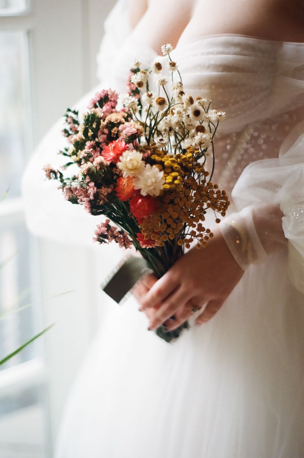 A delicate dried flower bouquet held against a white wedding dress in soft natural light.