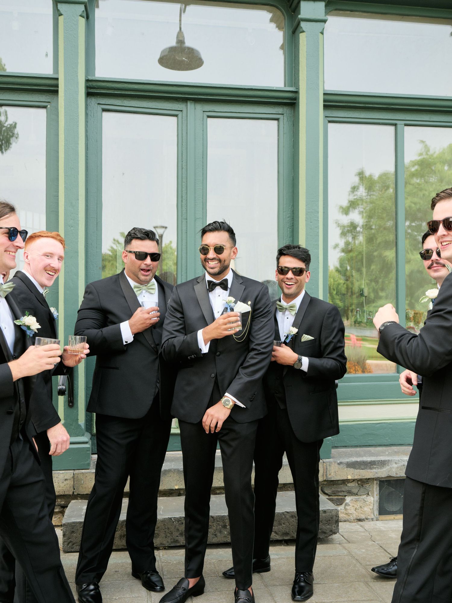 Groomsmen in black suits and sunglasses pose together in a fun lineup against a glass window exterior.