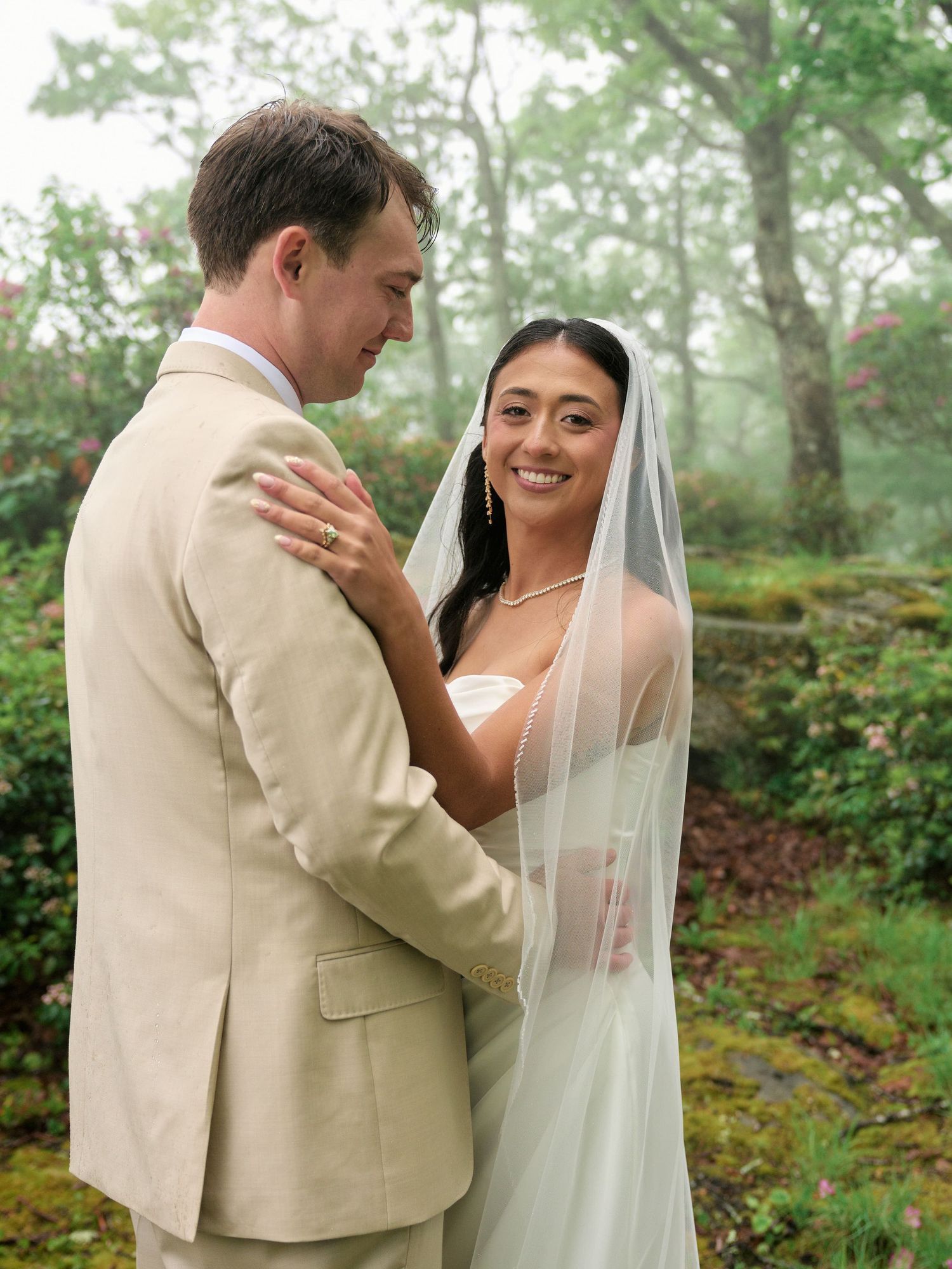 Bride in white gown and veil smiles at groom in tan suit during misty outdoor wedding ceremony.