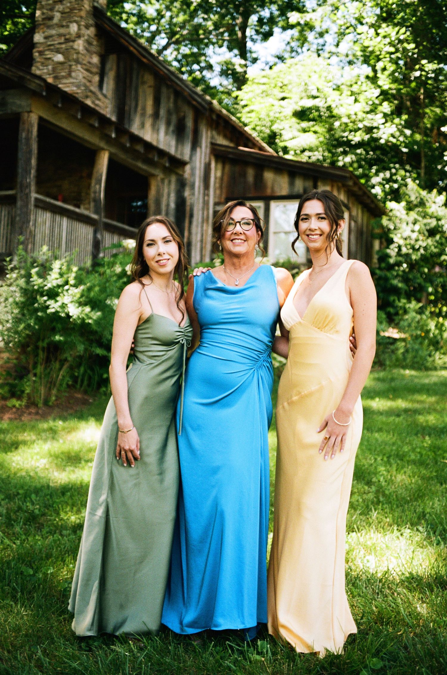 Three women in elegant floor-length gowns of sage, blue, and yellow pose together outdoors near a rustic barn.