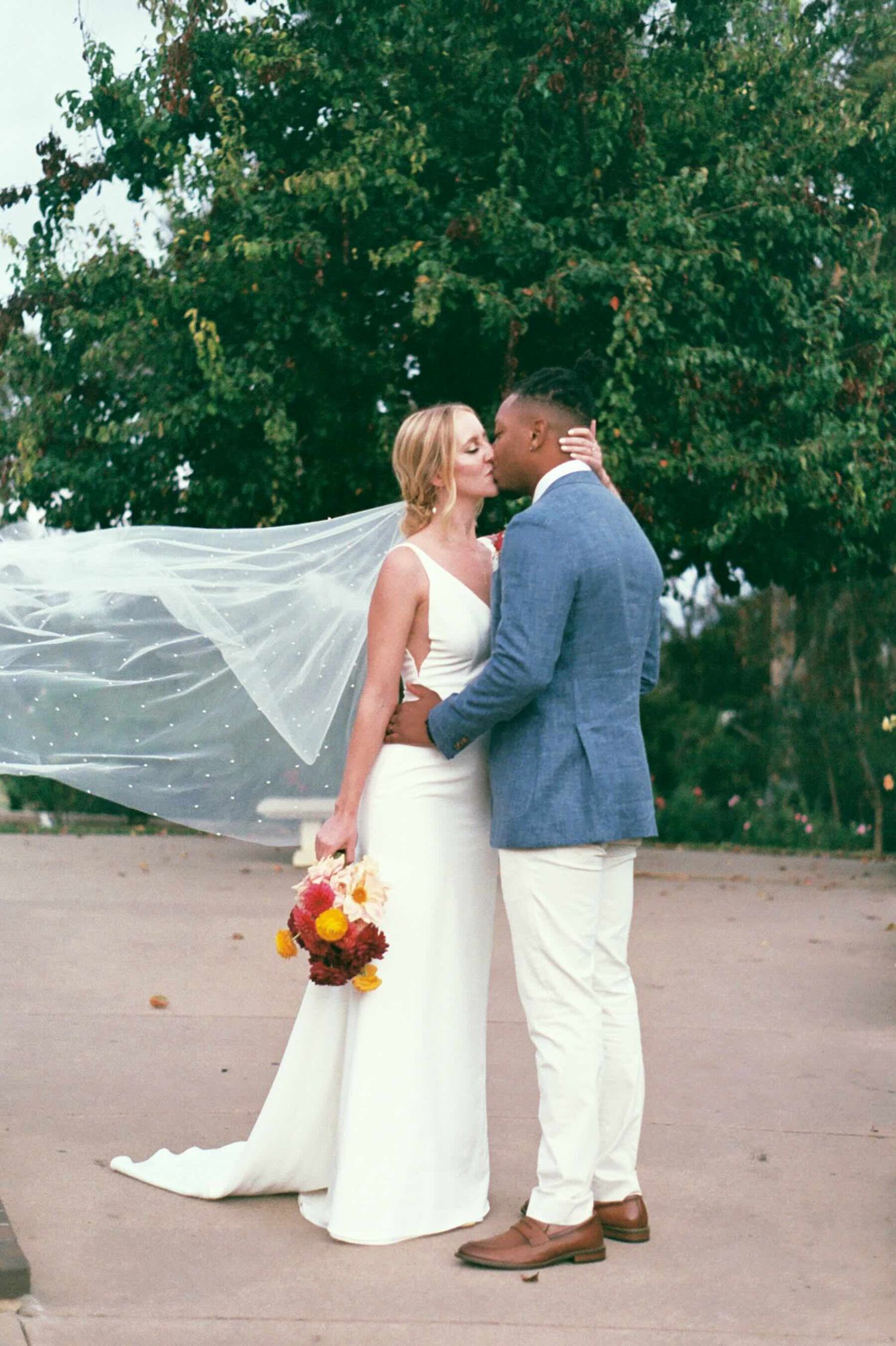 A romantic wedding couple embraces outdoors while the bride's veil flows dramatically in the wind.