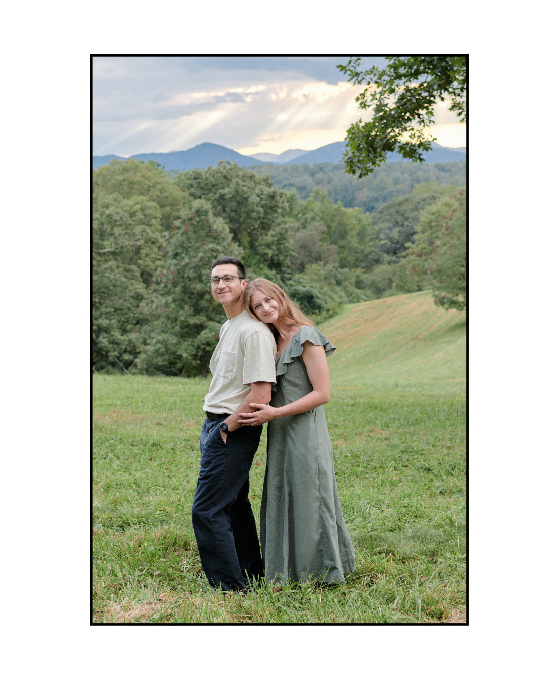 A couple stands together in a scenic mountain meadow with rolling hills and lush greenery in the background.