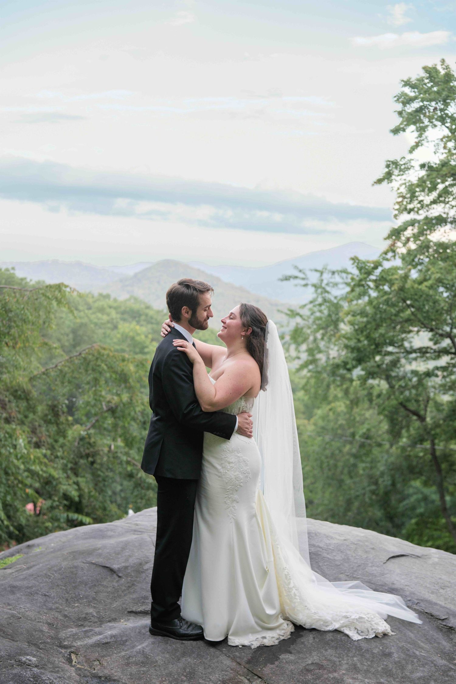 Bride and groom embrace on a rocky overlook with lush green mountains and cloudy sky backdrop at their outdoor wedding.