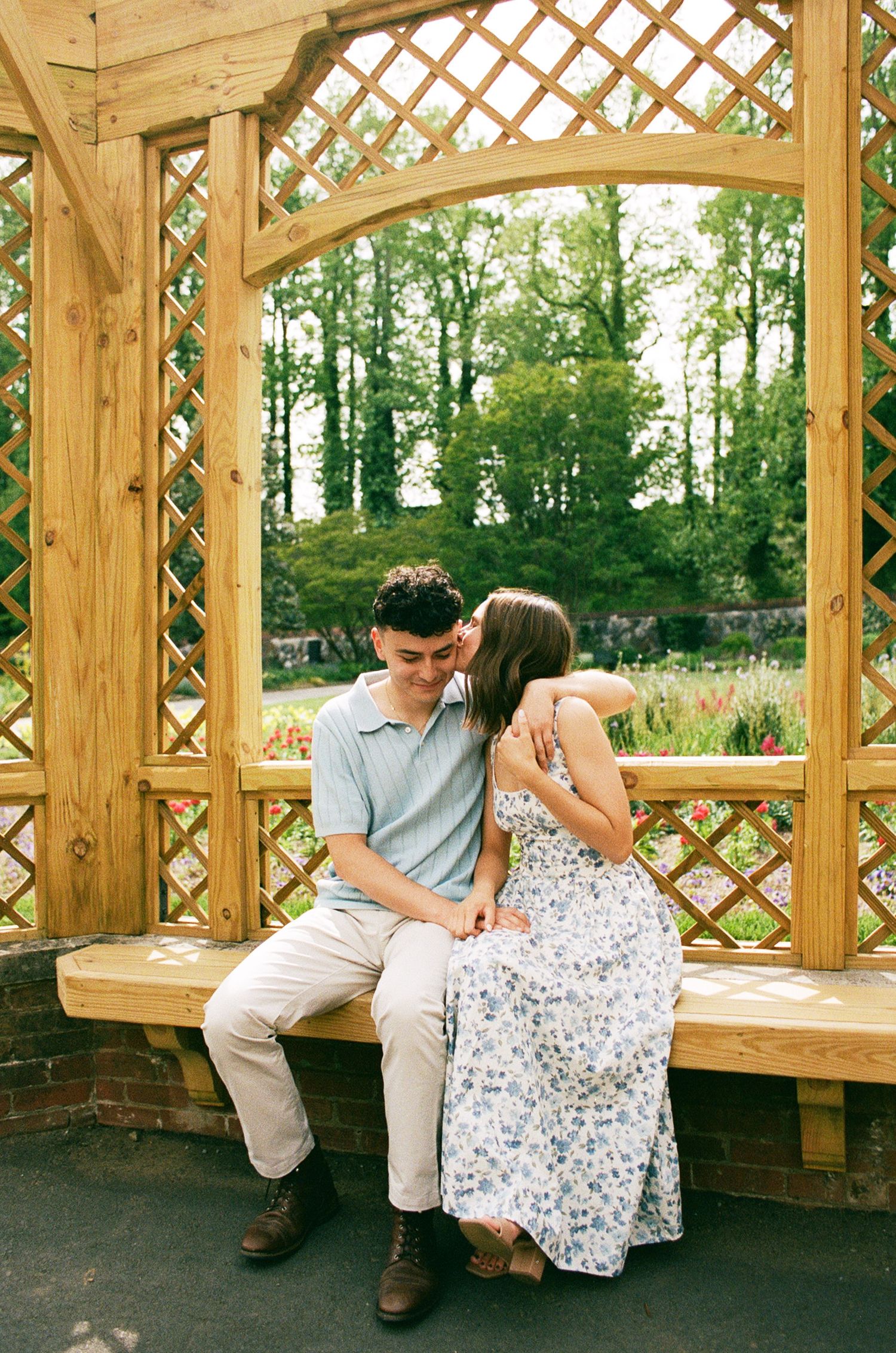 A couple shares a tender moment on a bench under a wooden garden gazebo, surrounded by lush green trees.