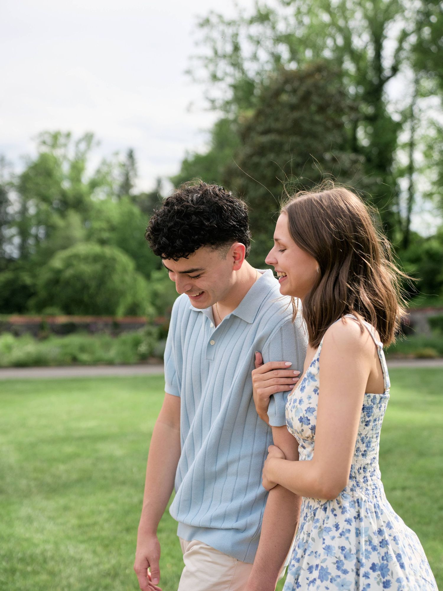A young couple laughs and walks together in a lush green garden, wearing casual summer attire.