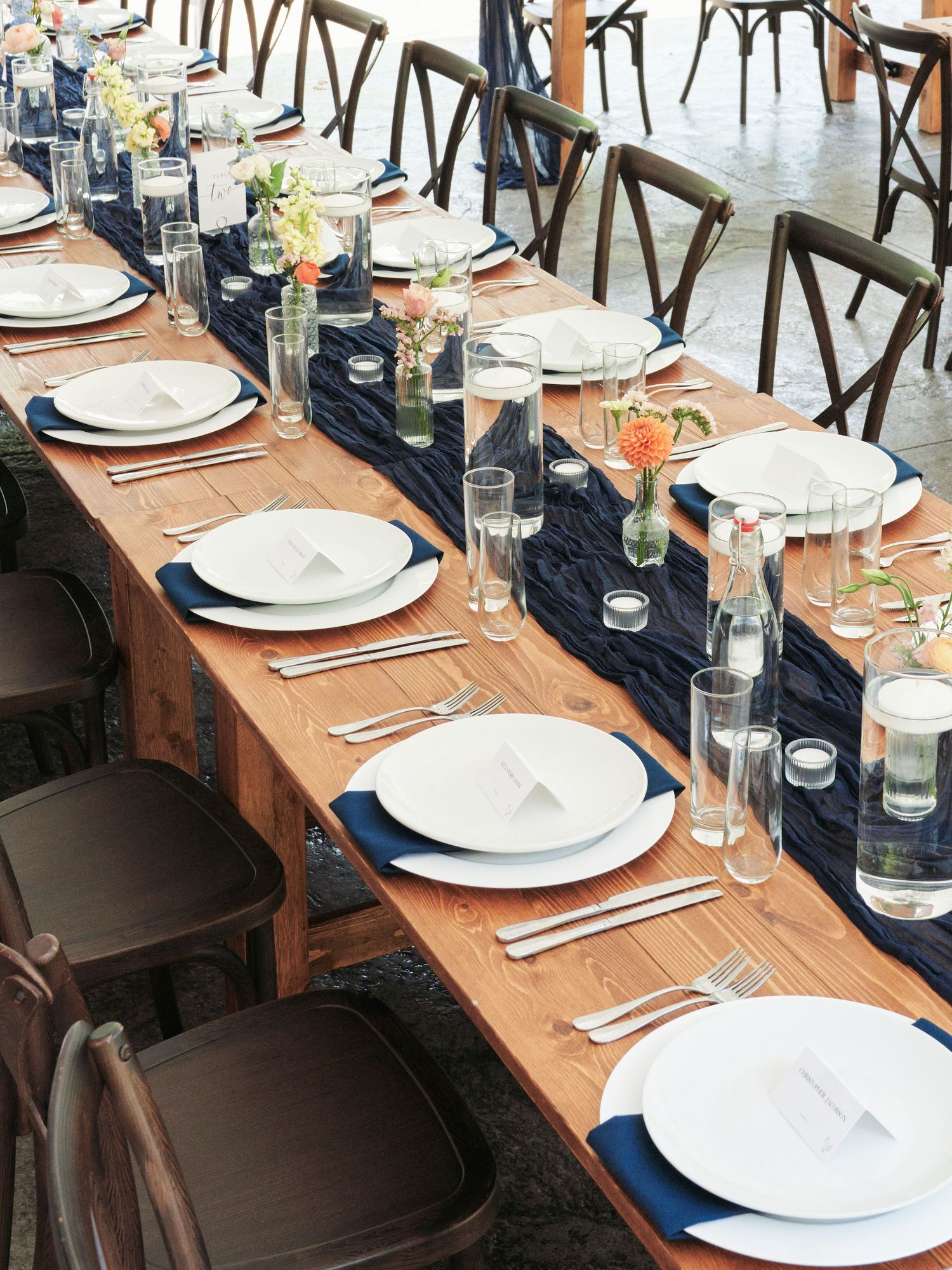 Elegant farm table wedding reception with navy blue runner, white plates, and floral centerpieces.