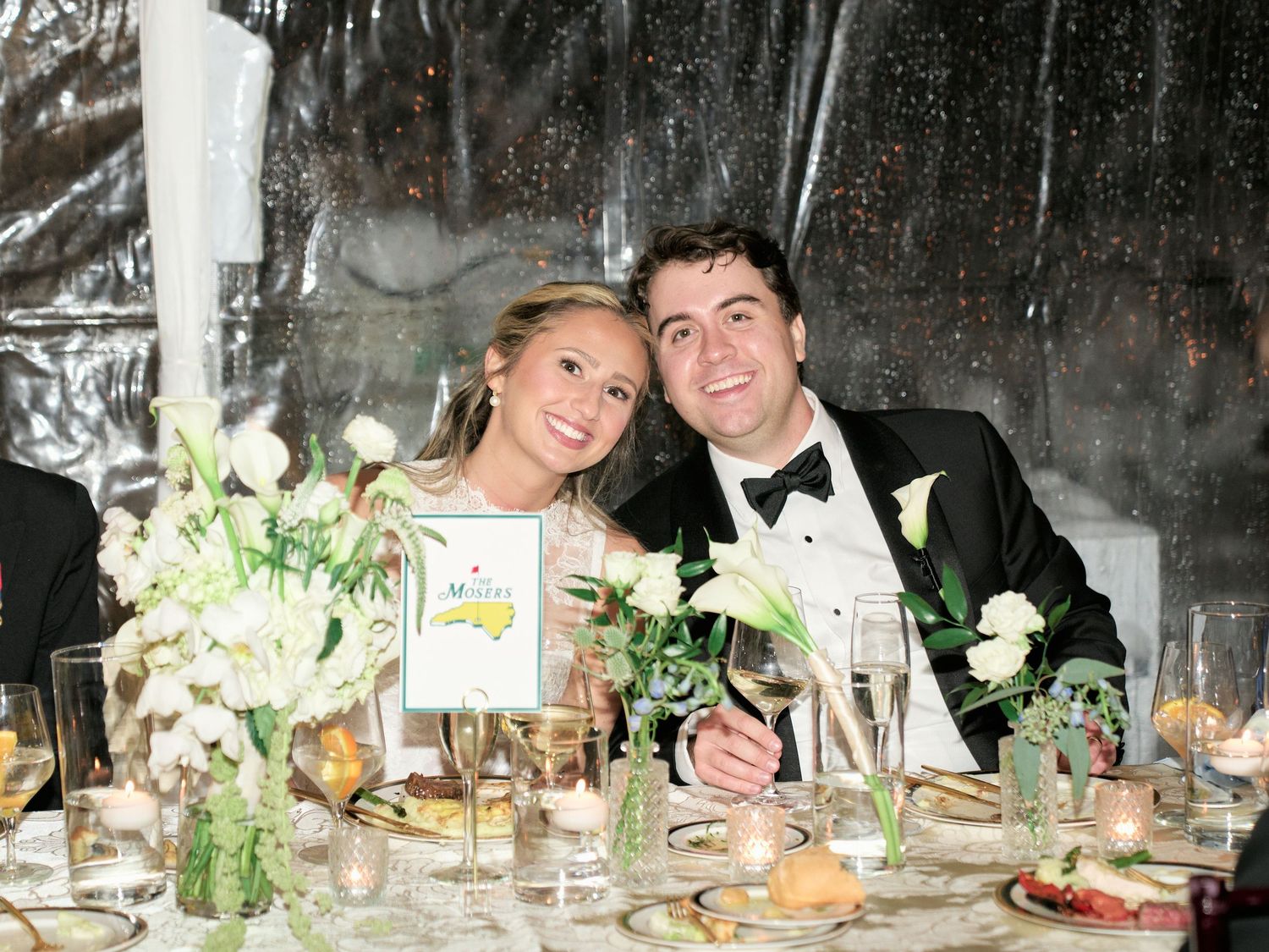 Smiling bride and groom seated at elegant wedding reception table with white floral centerpieces and candlelight.