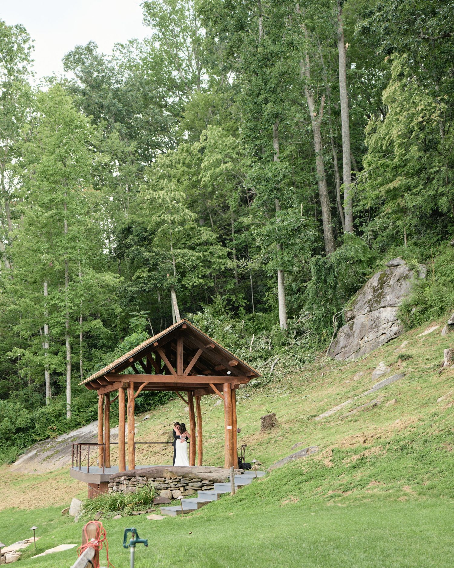 Couple sharing a kiss under a rustic wooden gazebo surrounded by lush green forest and rocky hillside.