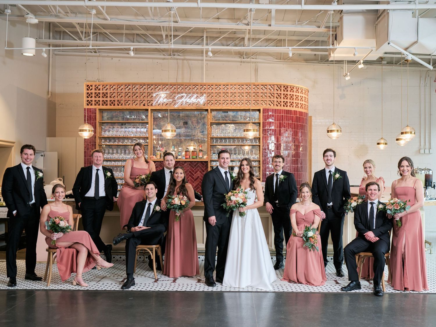 Wedding party poses in dusty rose gowns and black suits inside an elegant venue with a decorative bar backdrop.