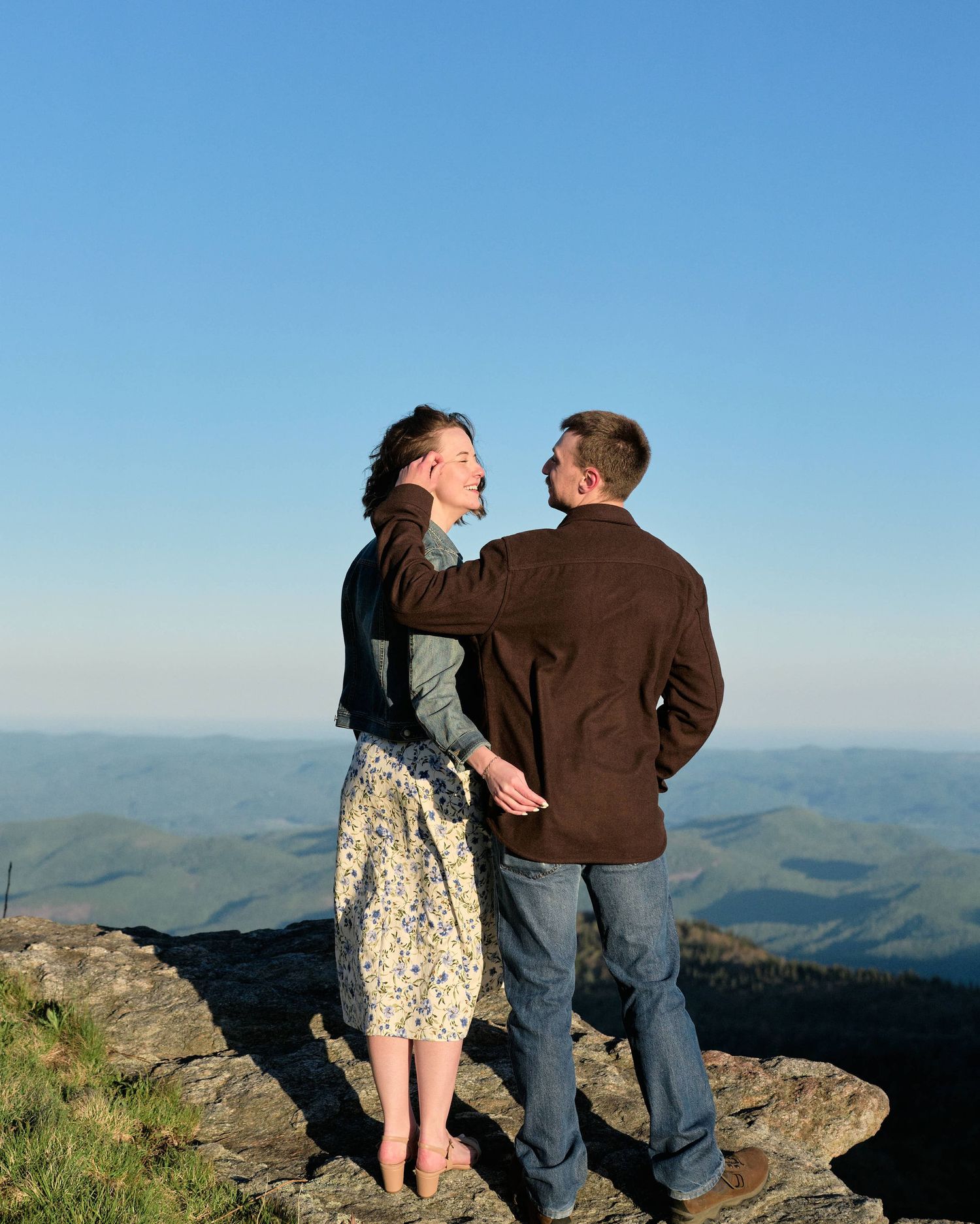 Couple standing on rocky mountain summit, embracing with scenic blue ridge mountain range and clear sky backdrop.