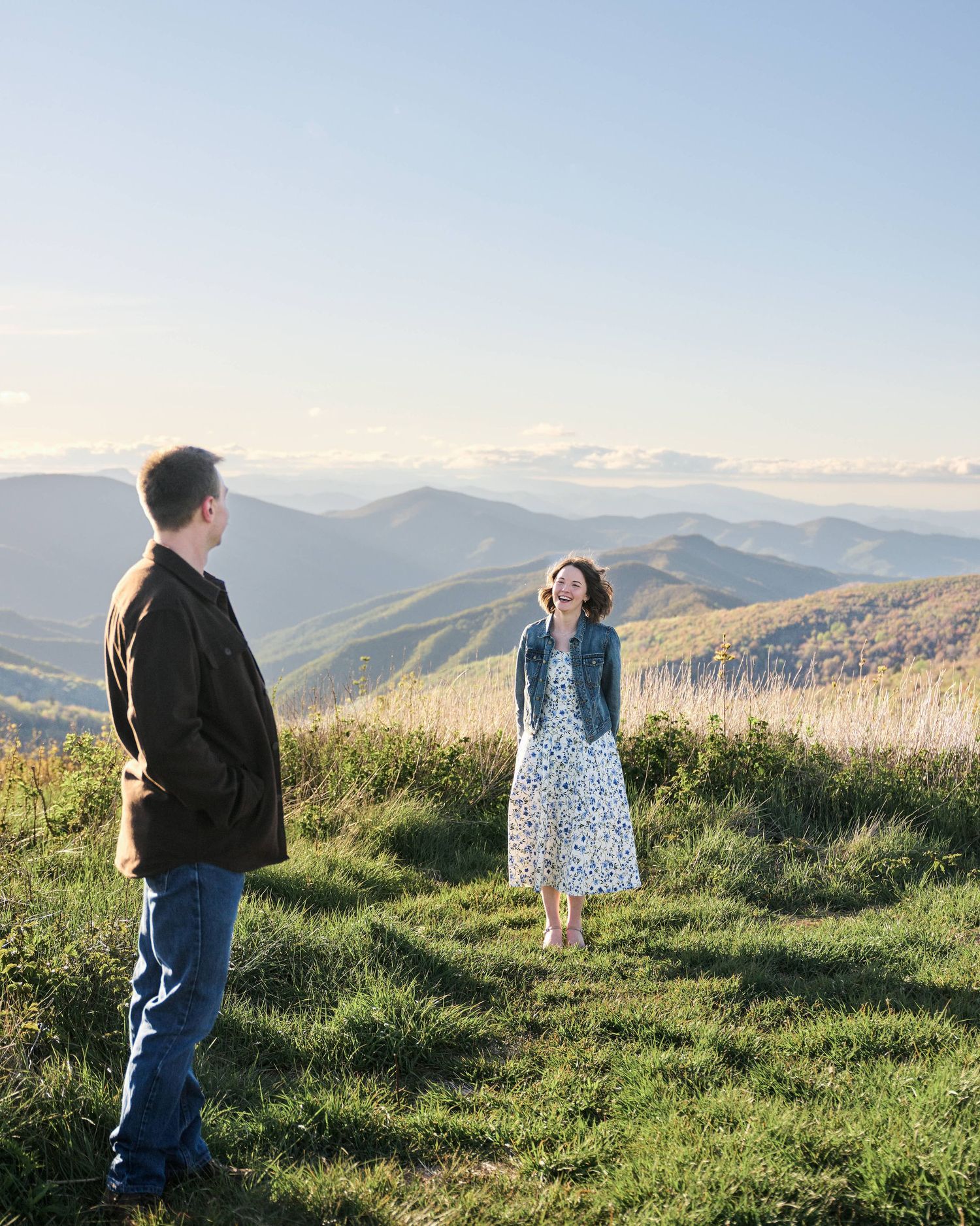 Couple laughing together on a scenic mountain overlook with rolling blue ridges and clear sky backdrop.
