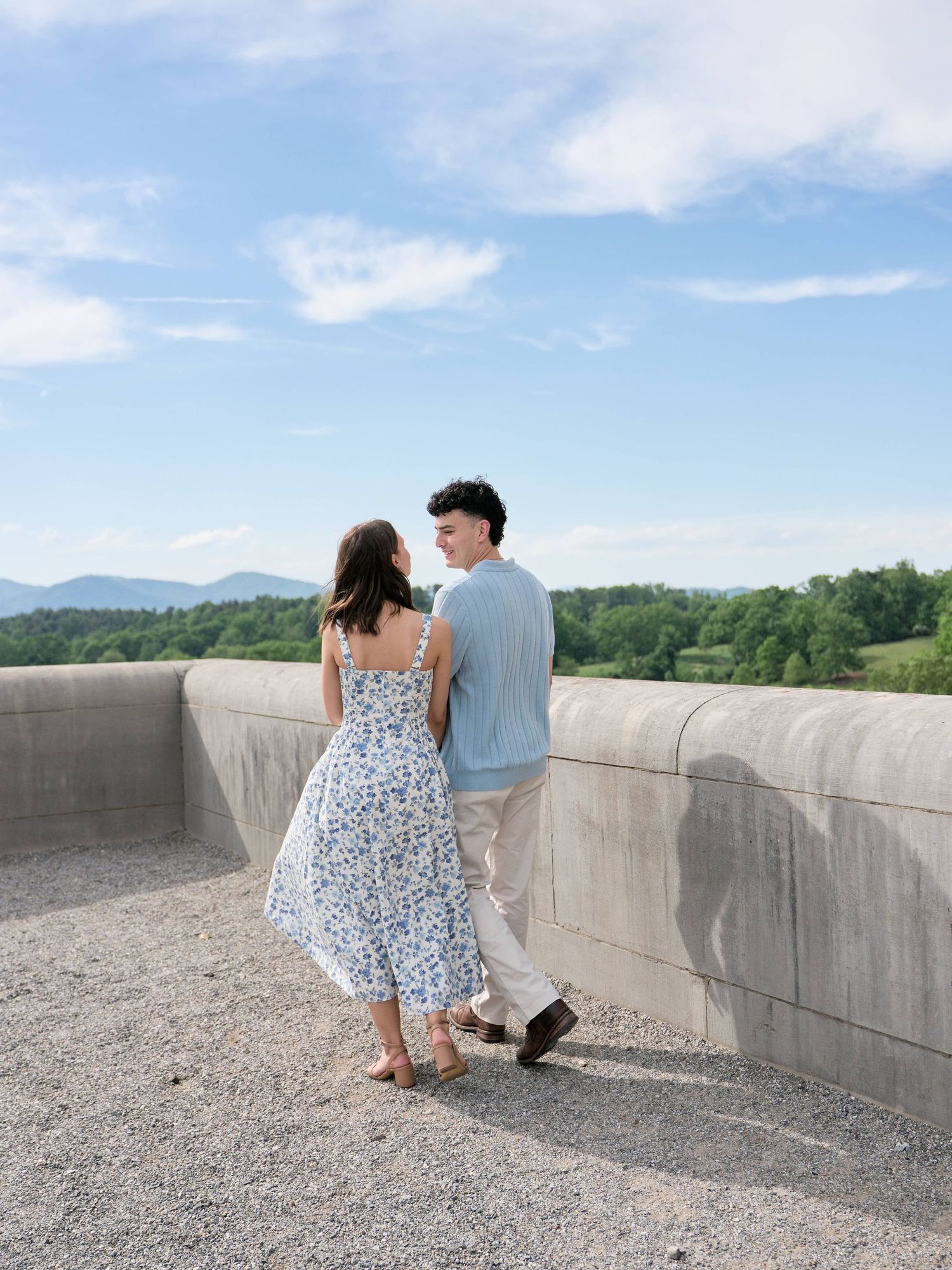 A couple in summer attire stands on a concrete overlook, gazing at each other with green hills and blue sky behind them.