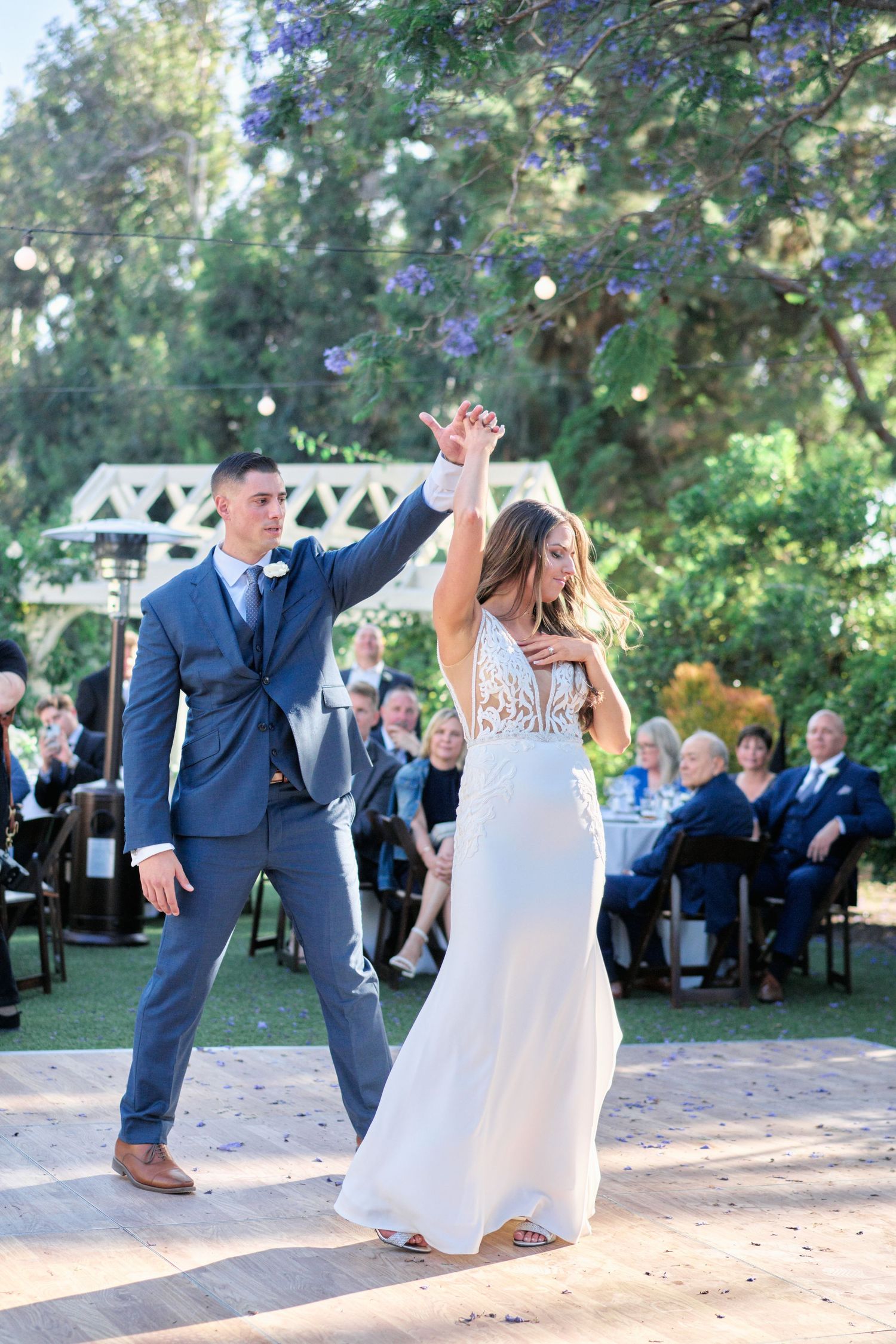 Newlyweds share their first dance outdoors with guests watching in a garden setting.