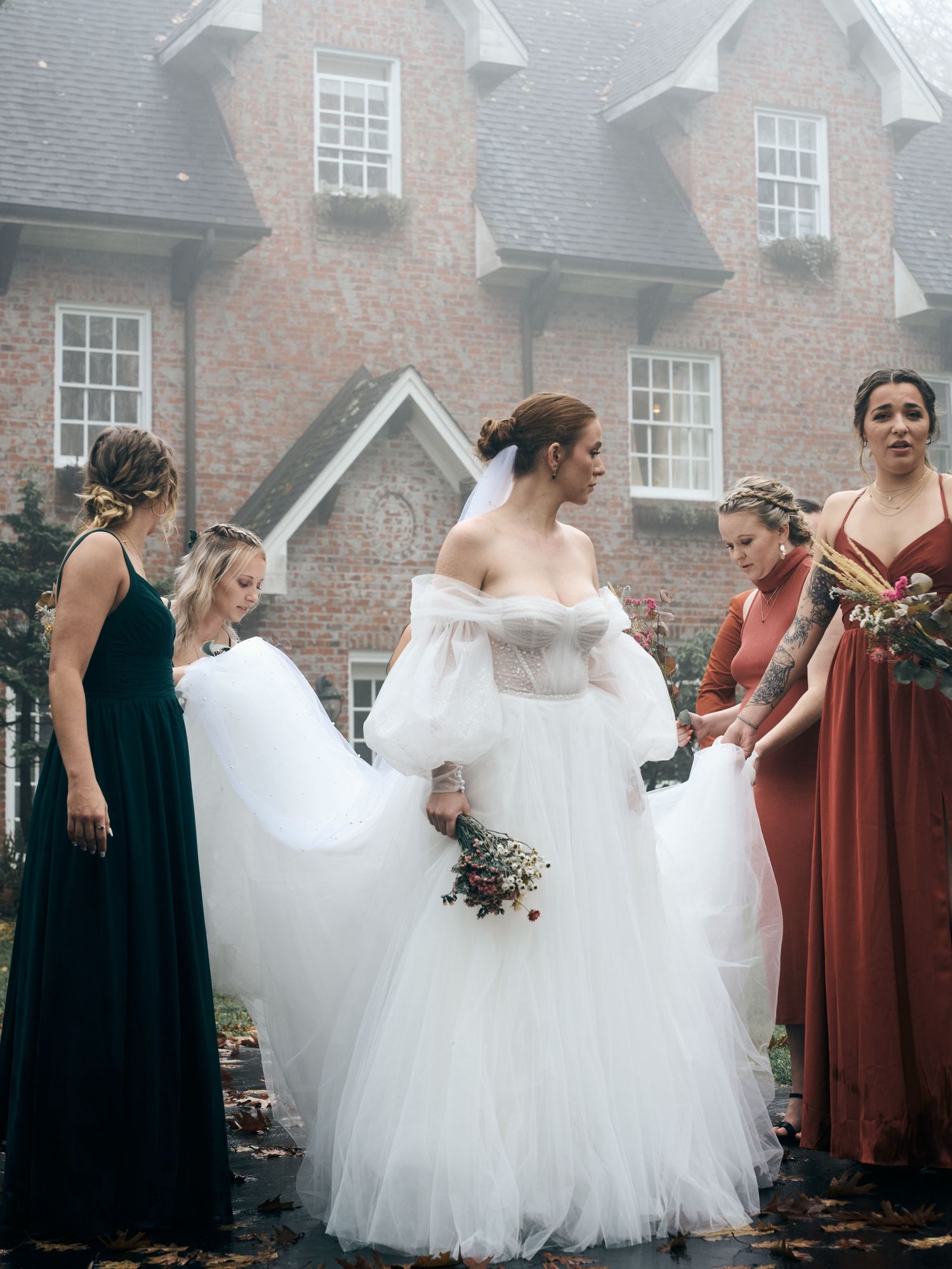 A wedding party helps adjust a bride's flowing white gown in front of a brick mansion.