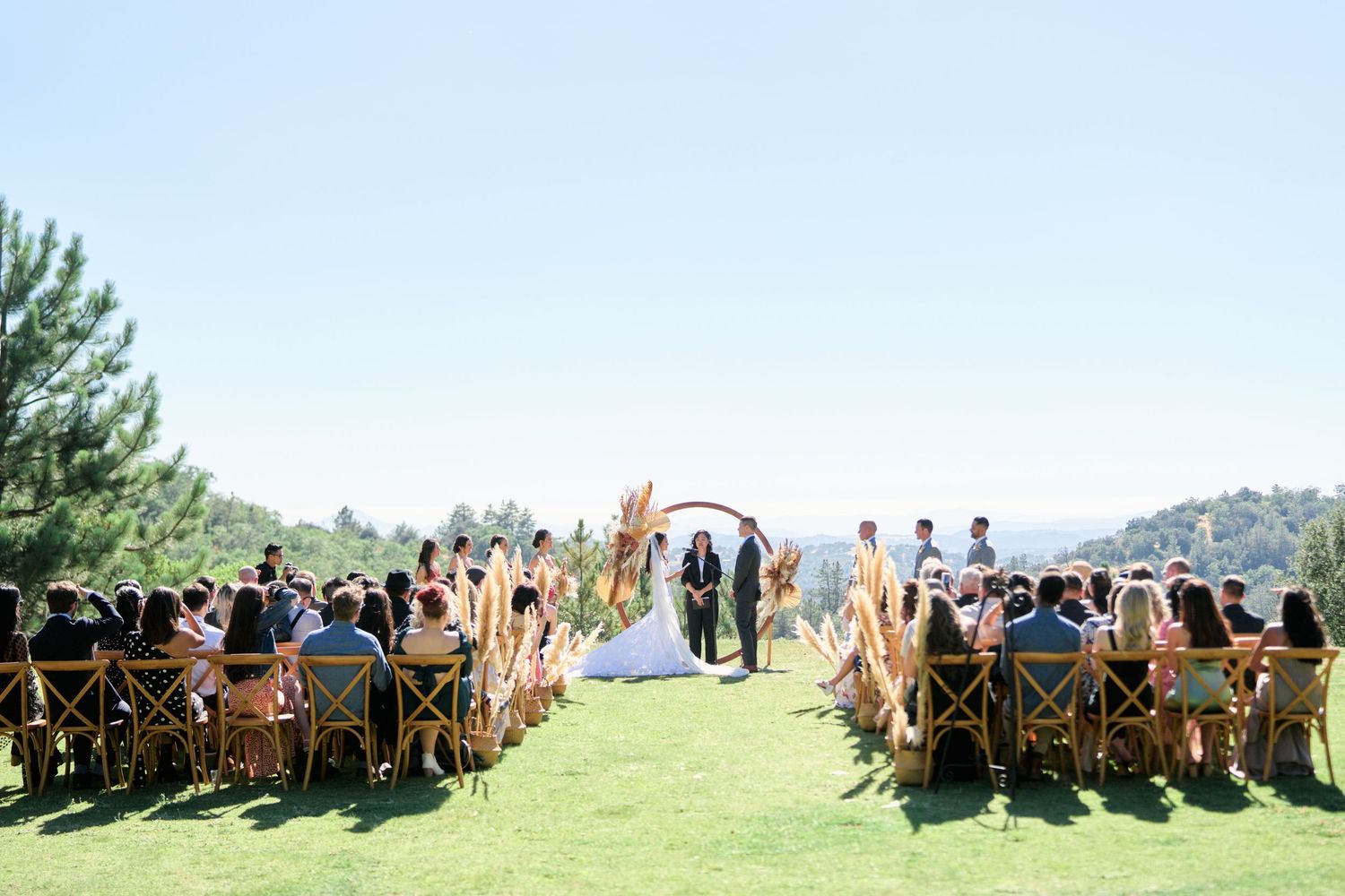 Outdoor wedding ceremony on a grassy hilltop with wooden chairs arranged in rows for guests.