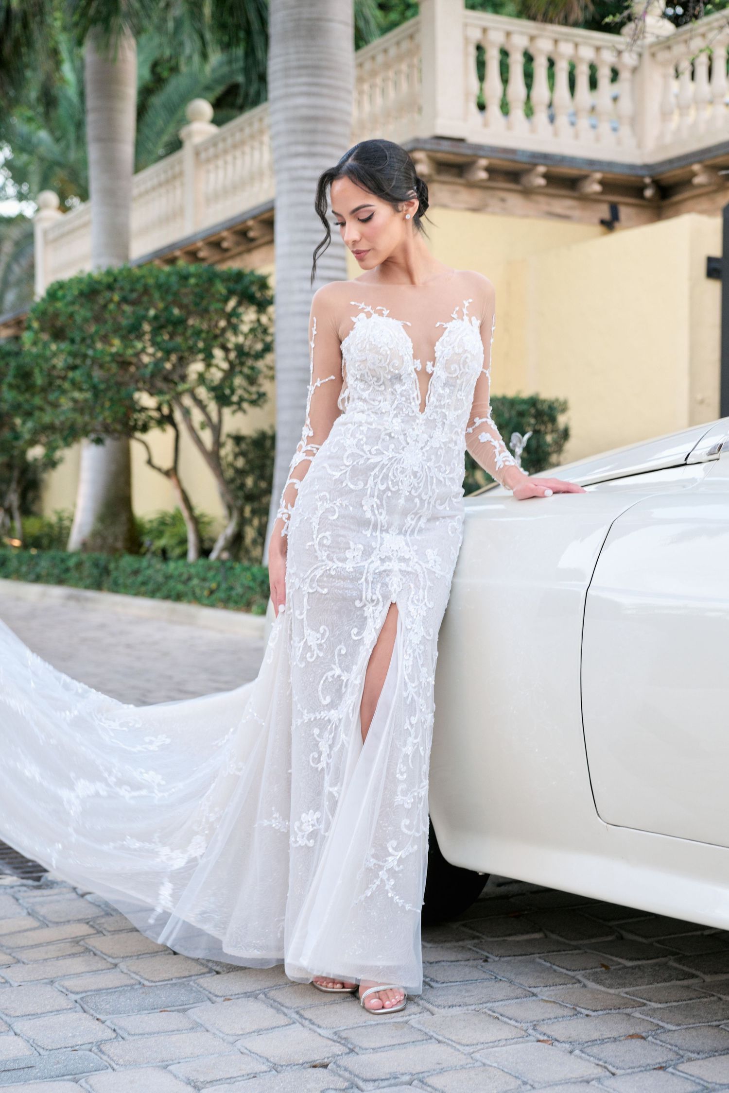 Full length view of bride in sheer lace wedding gown with side slit beside vintage car.