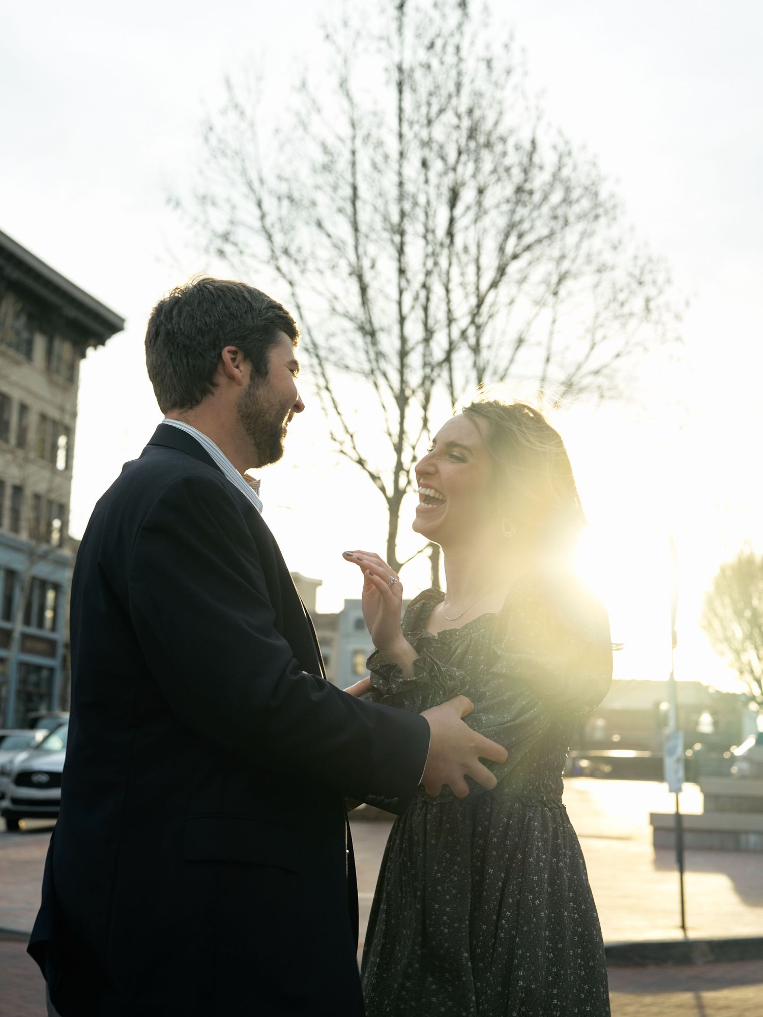 A couple laughing together in warm sunlight with a building and trees in the background.