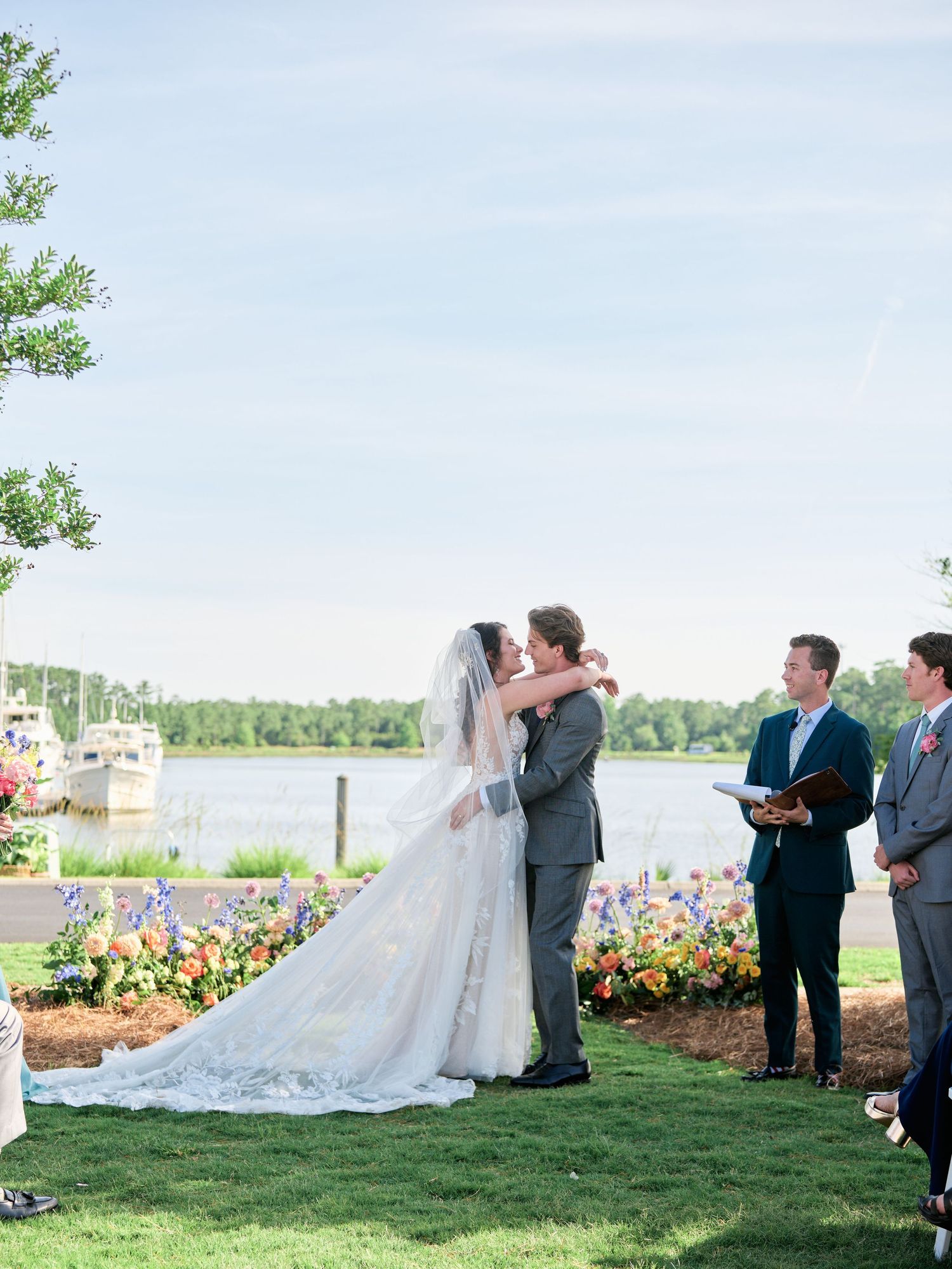 Outdoor wedding ceremony takes place by a lake with colorful flower garden and scenic water view in background.