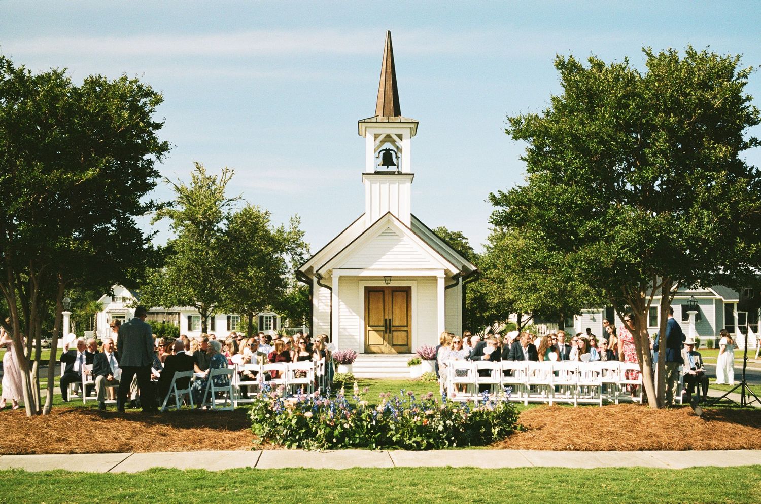 A charming white chapel with tall steeple stands surrounded by guests at an outdoor wedding ceremony.