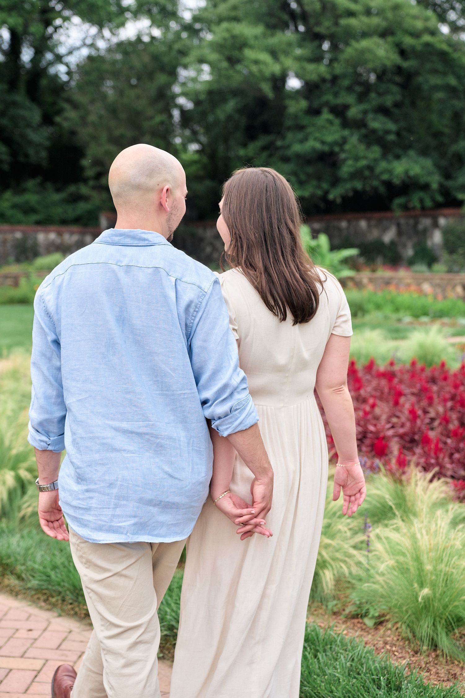 Couple walks hand in hand on brick path through garden with red flowers.