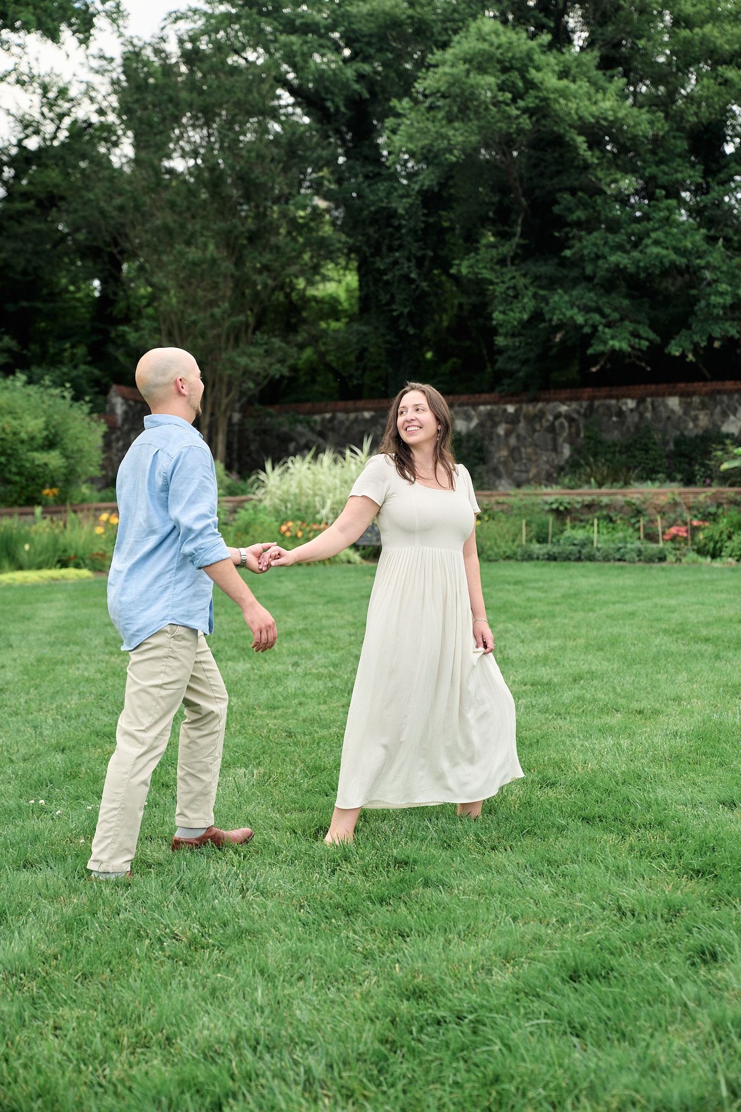 A couple dances playfully on a green lawn with gardens and stone walls in the background.