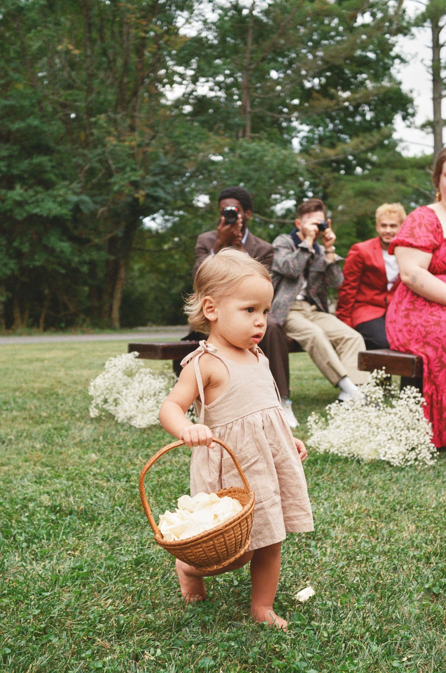 Young flower girl in beige dress carries basket across lawn during outdoor wedding ceremony.
