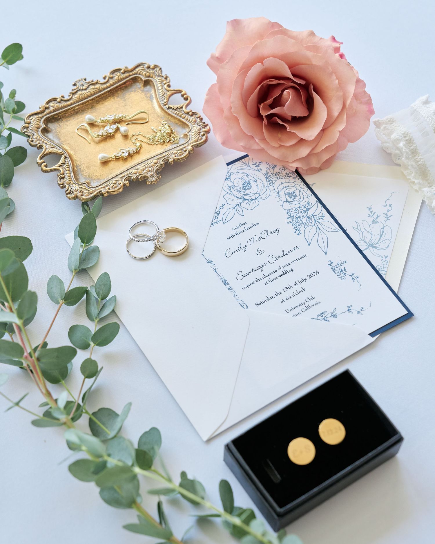 Wedding details including invitation, rings, earrings, and pink rose on white background.