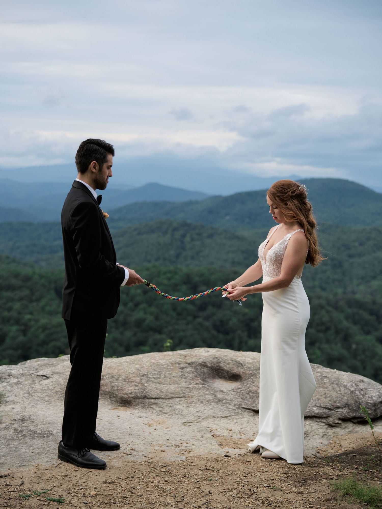 A couple in wedding attire stands on a mountain overlook with a dramatic view of rolling hills and cloudy skies.