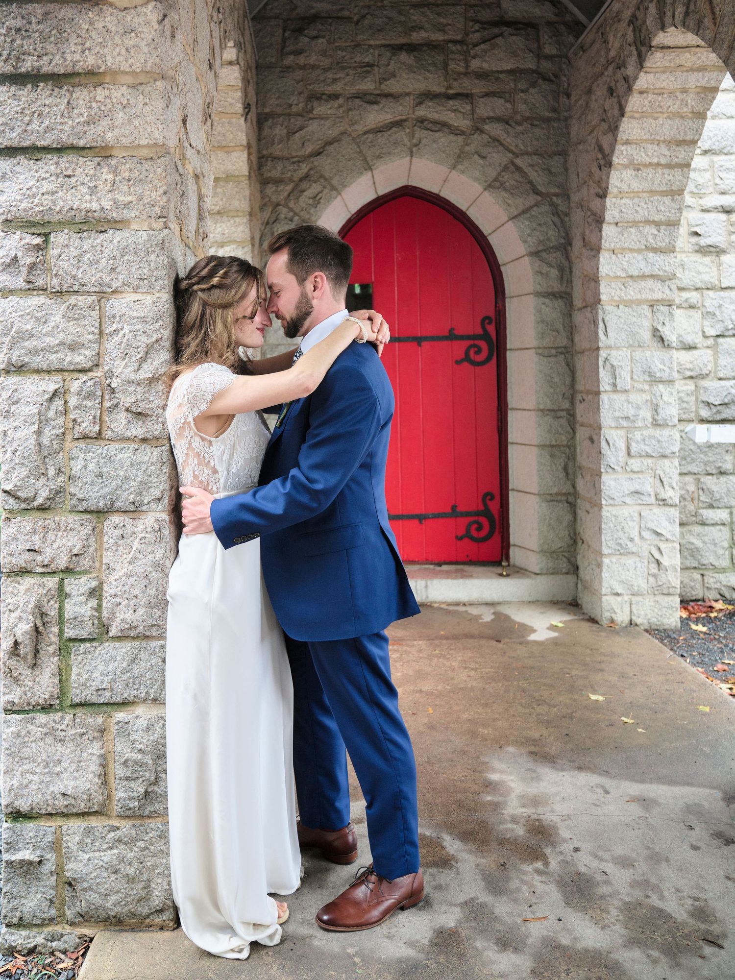 A romantic moment between newlyweds embracing in front of a stone church entrance with a vibrant red door.