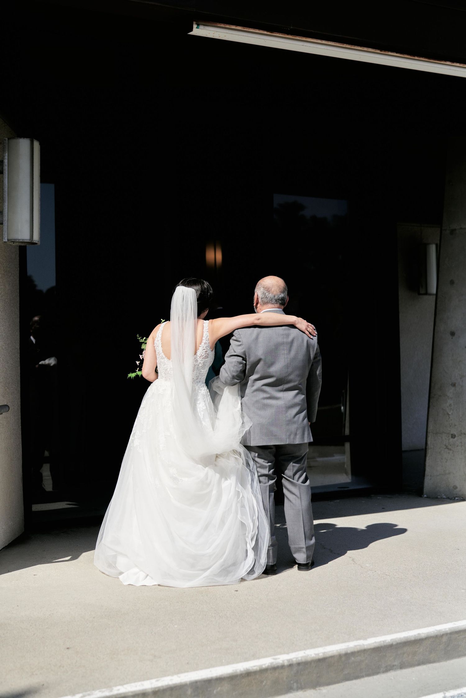 A bride in white gown walks arm in arm with an elderly man in grey suit entering a building.