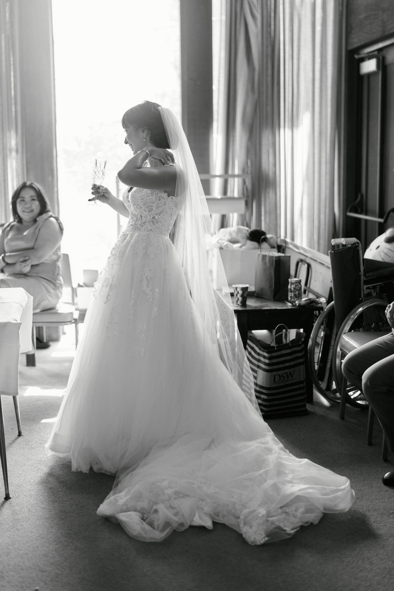 A bride in a flowing white gown and veil stands in a sunlit room during wedding preparations.